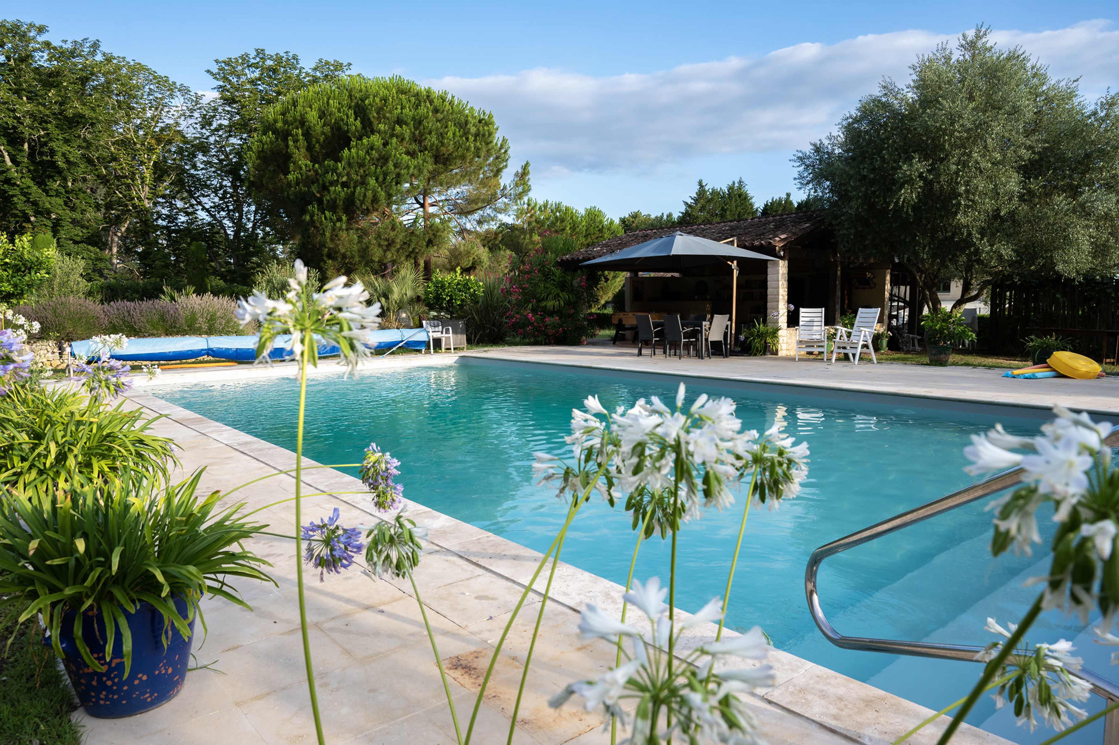 A clear blue swimming pool is surrounded by lush greenery and flowers, with a shaded seating area nearby.