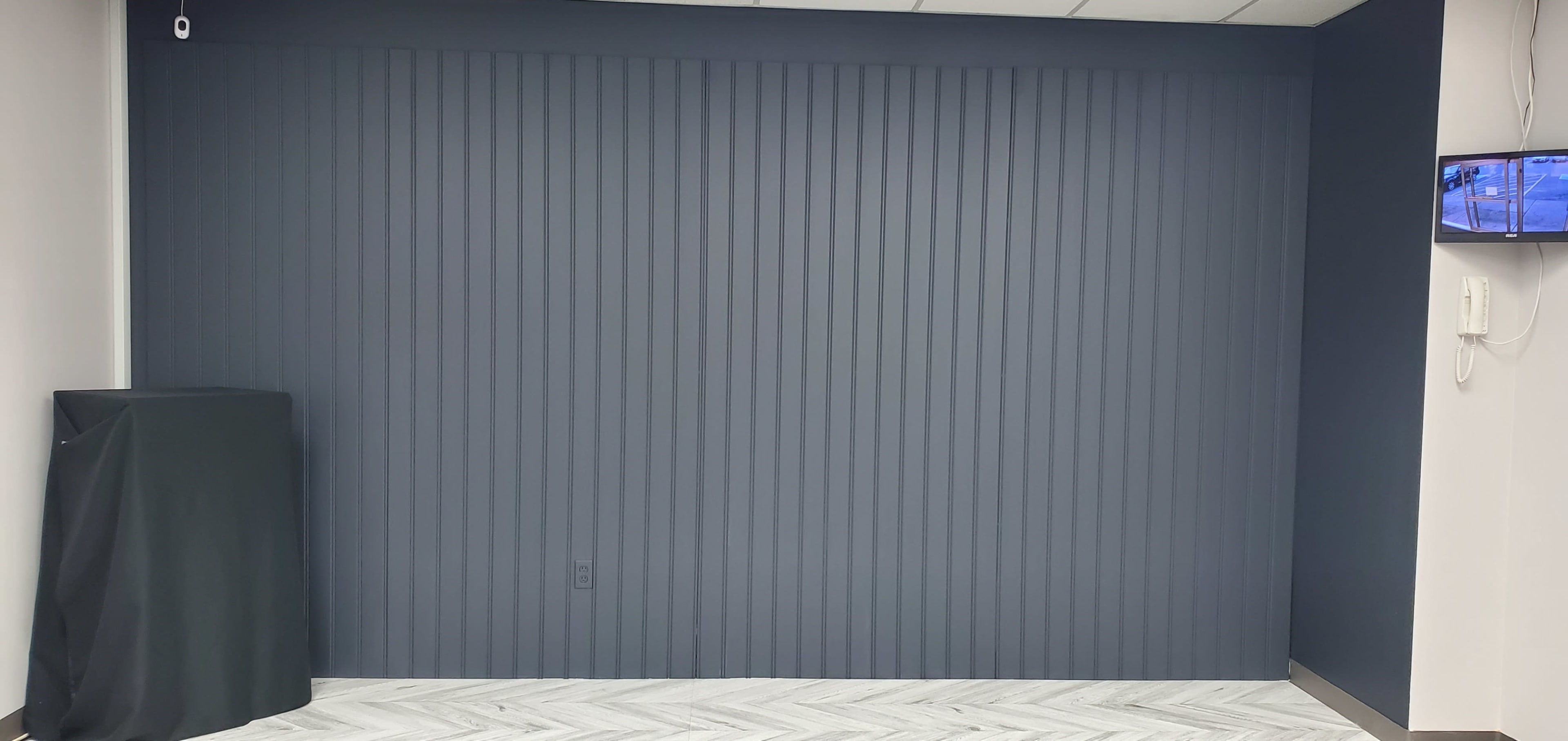 A room with a vertical striped dark gray wall, a light-colored herringbone floor, and a black object covered with a cloth in the corner.