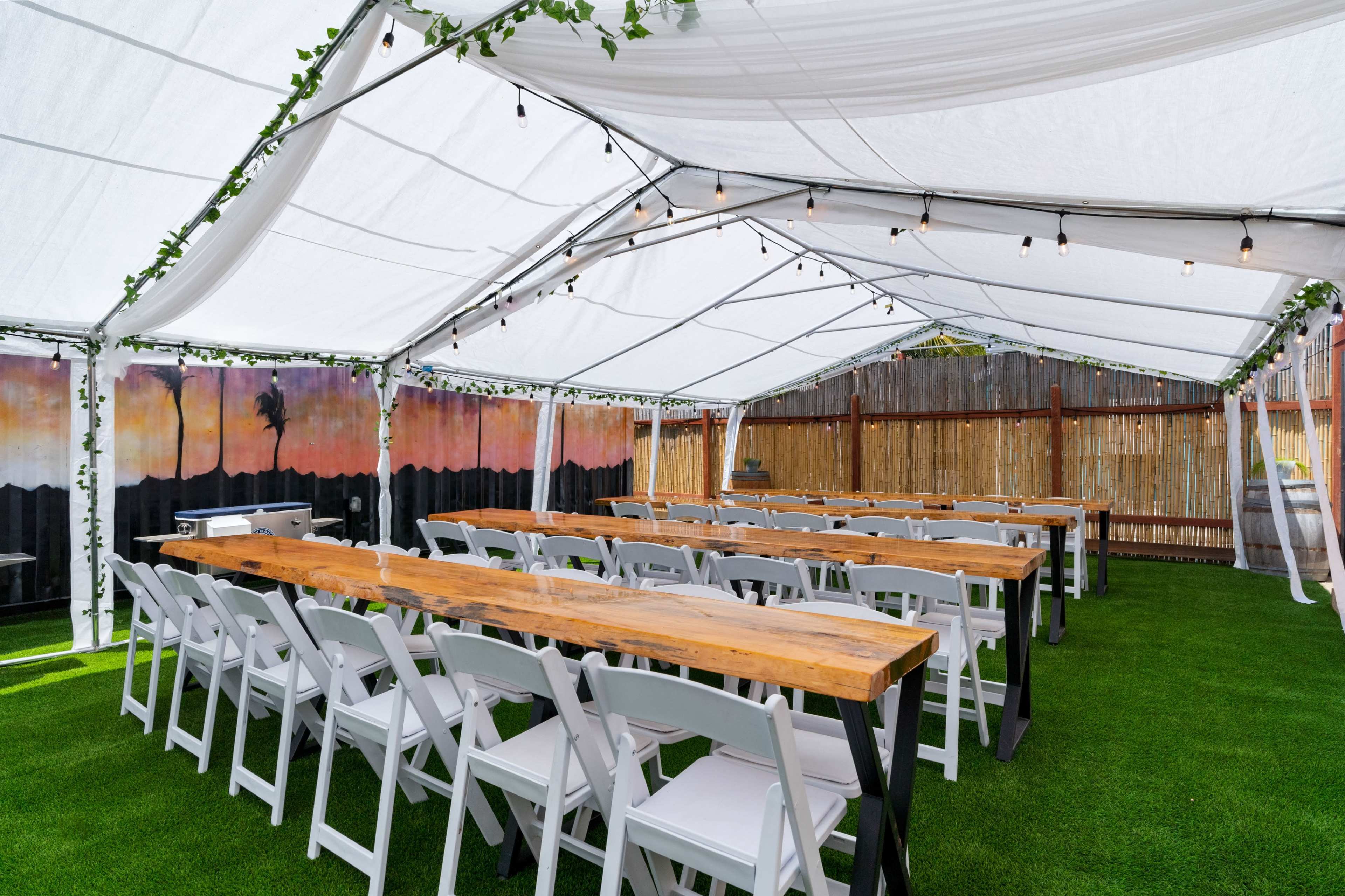 A tented outdoor venue set up with long wooden tables and white chairs arranged on artificial grass, decorated with greenery and string lights.