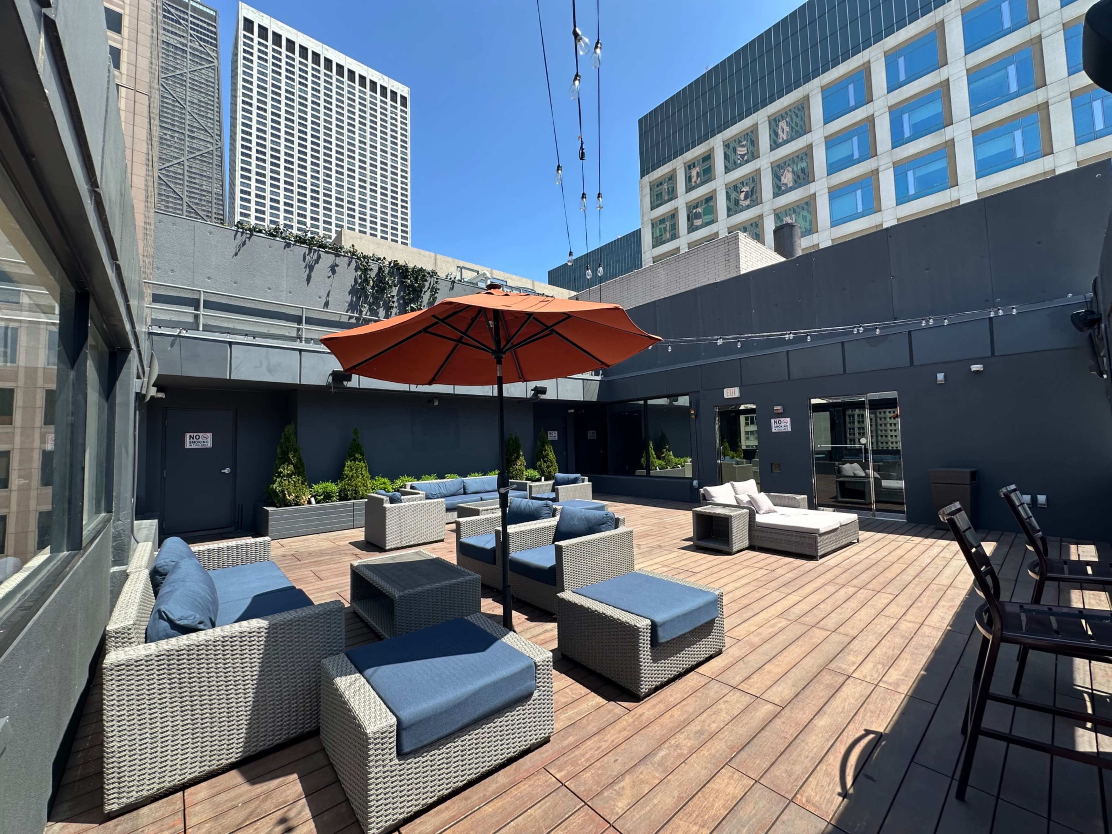 The image shows a spacious rooftop terrace with several seating areas, an orange umbrella, and a view of nearby buildings under a clear blue sky.