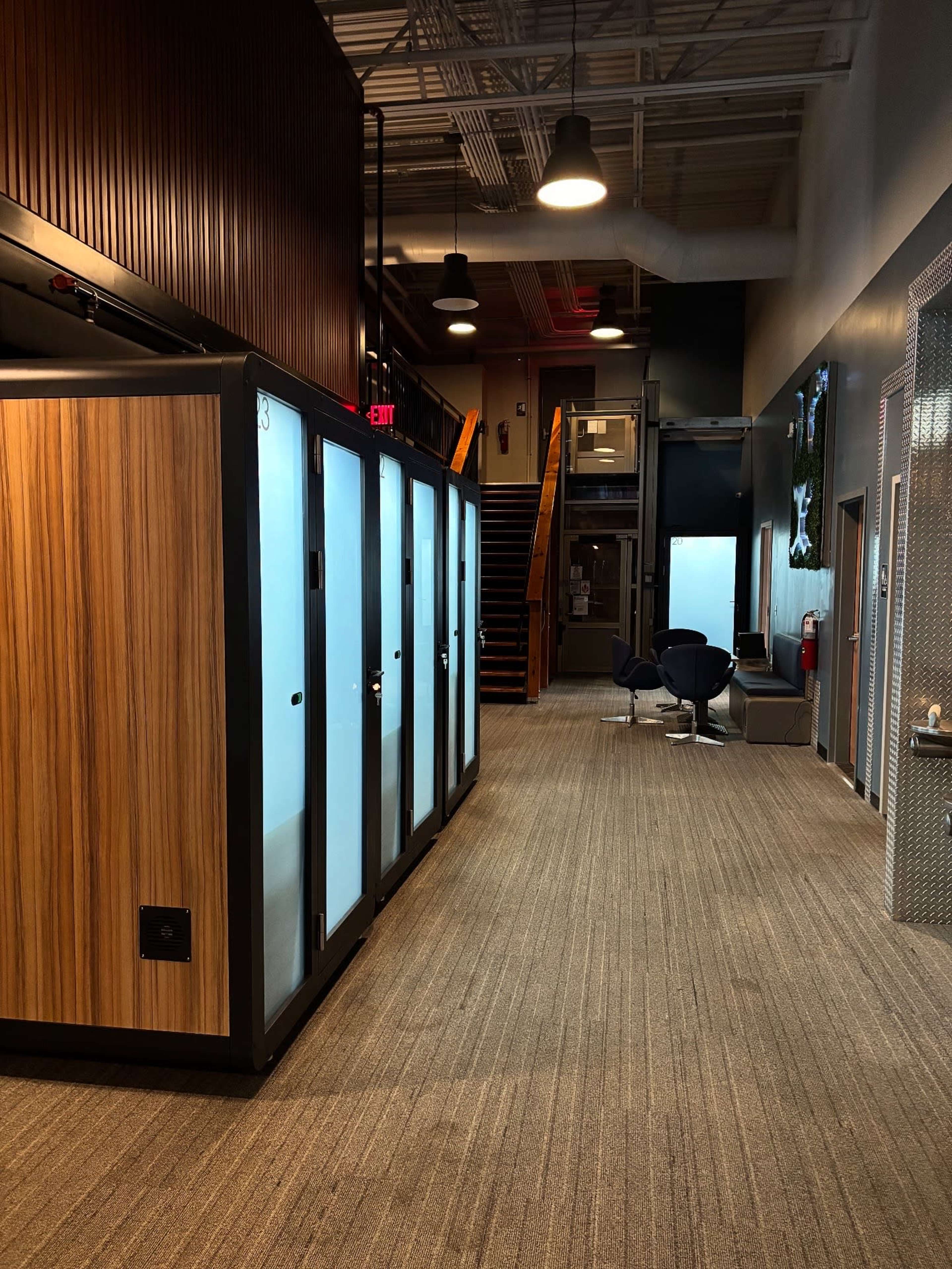 The image shows a hallway in an office space featuring several private phone booths on one side and a staircase leading to another level on the opposite side.