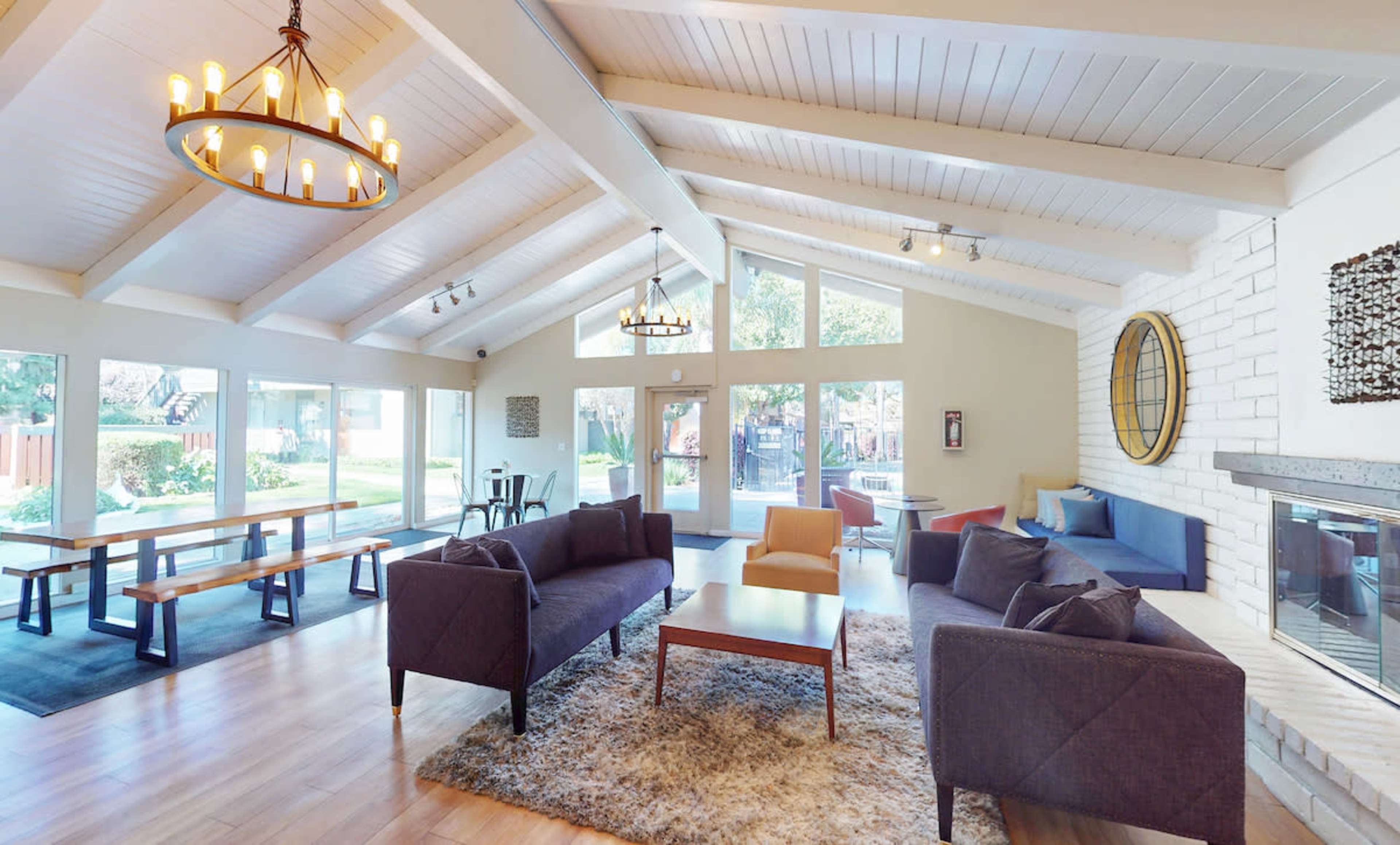 The image shows a spacious living area with high ceilings, large windows, and modern furniture arranged around a central coffee table.