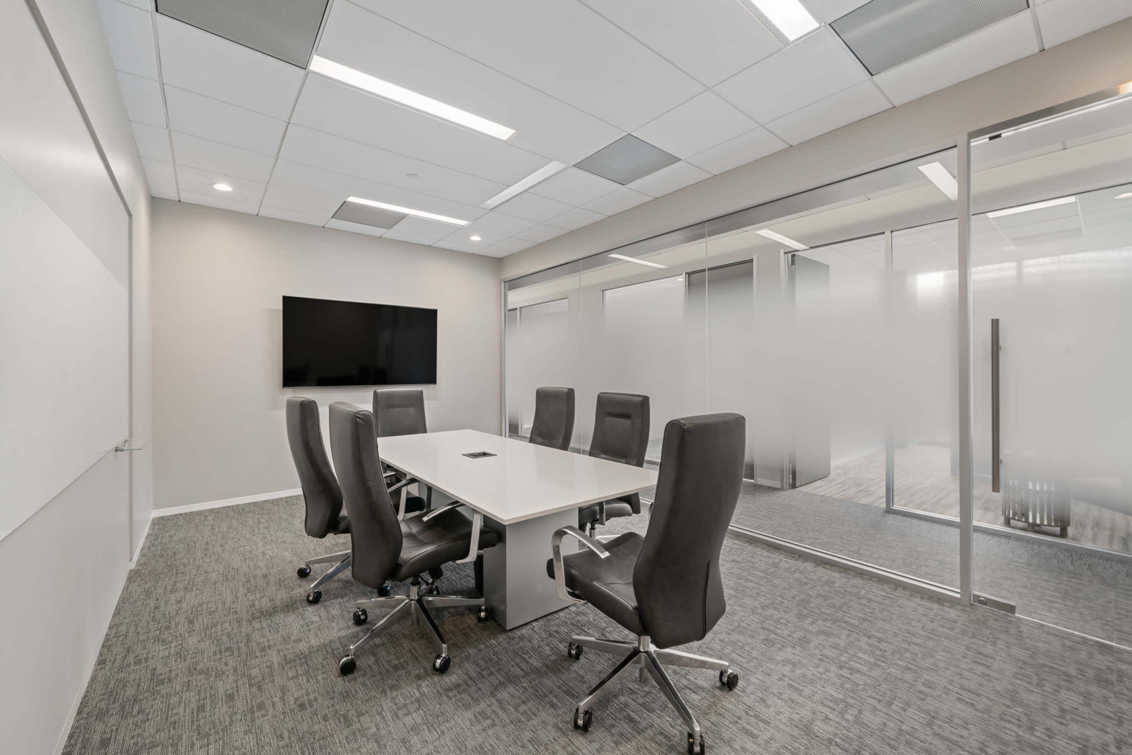 The image shows a modern conference room featuring a long white table surrounded by six ergonomic chairs, with a large screen mounted on the wall and glass partitions.