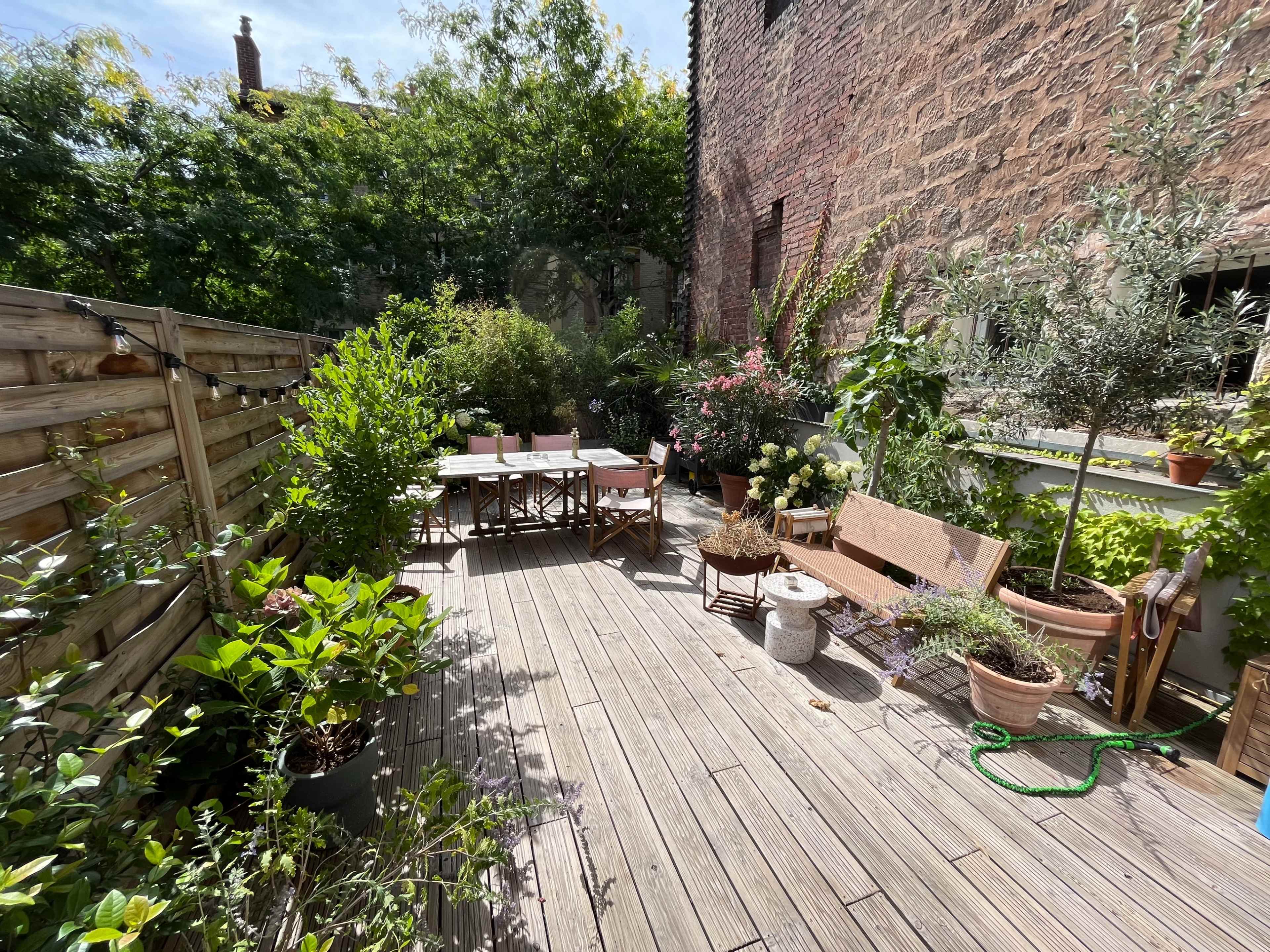 A wooden deck surrounded by greenery, with a dining table set up and various potted plants arranged around the area.