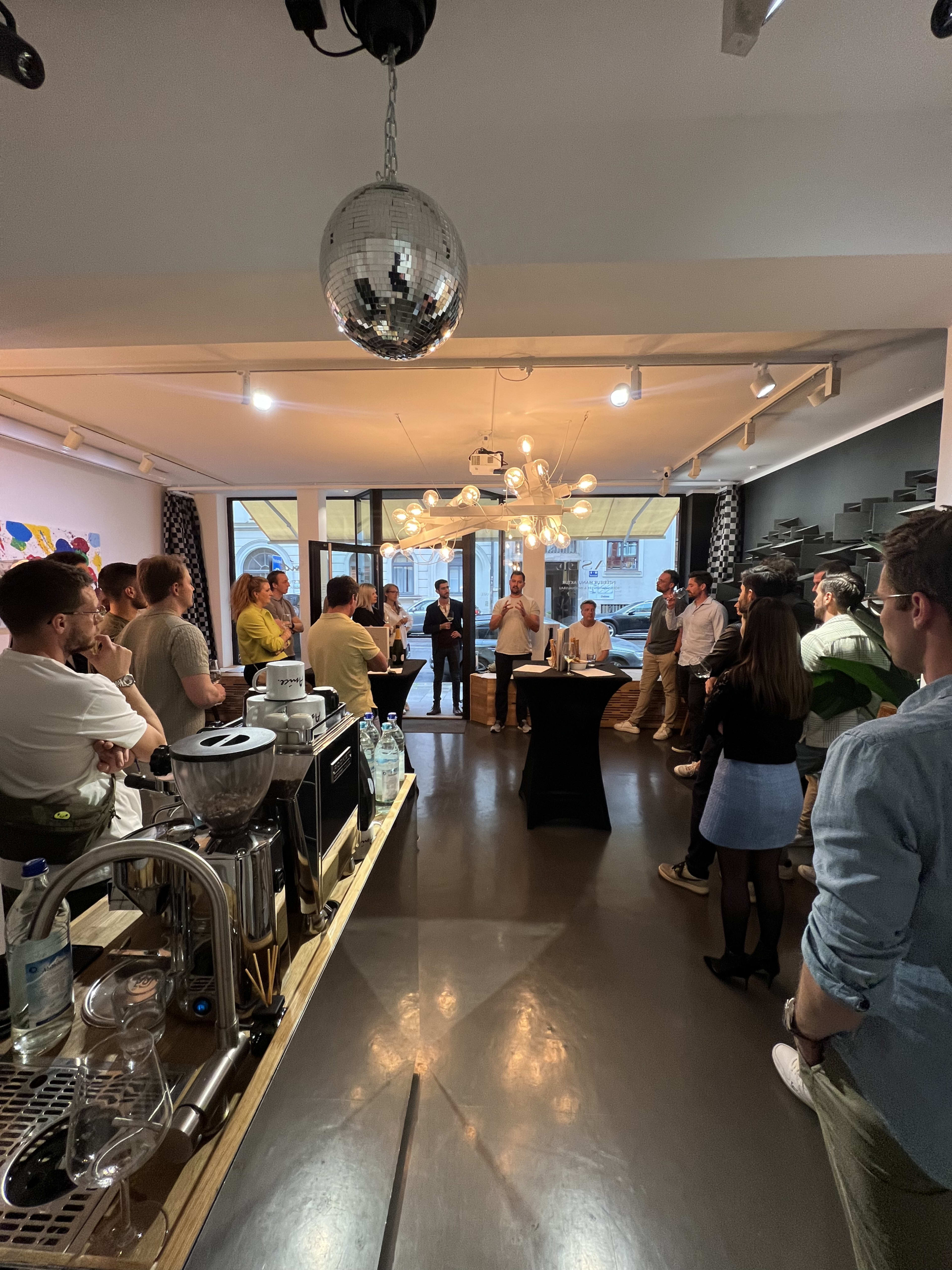 A group of people stands in a brightly lit café, gathered around a speaker at a black table while others observe from the surrounding area.