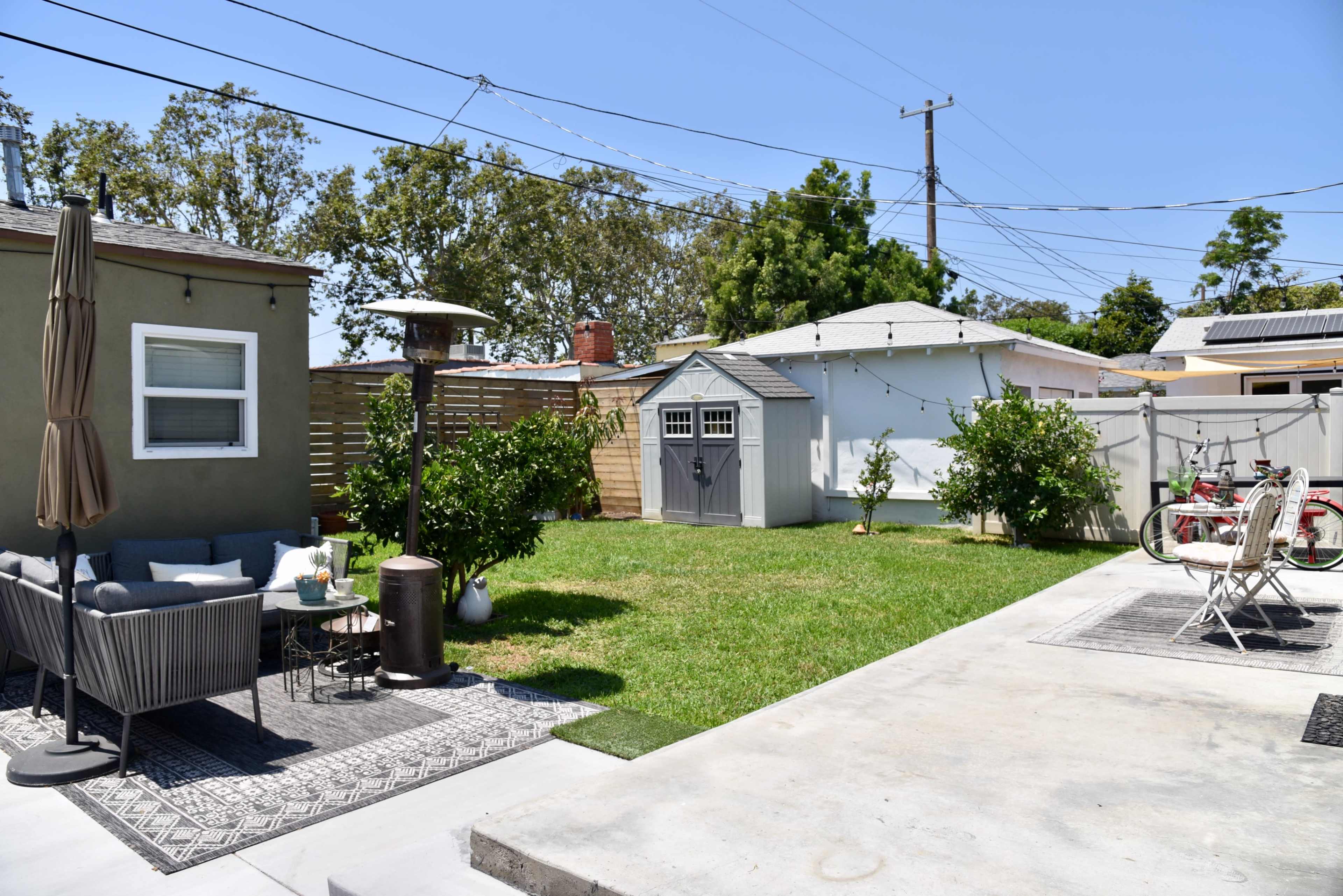 A backyard with a seating area, a small shed, and a grassy space bordered by fences and trees under a clear blue sky.