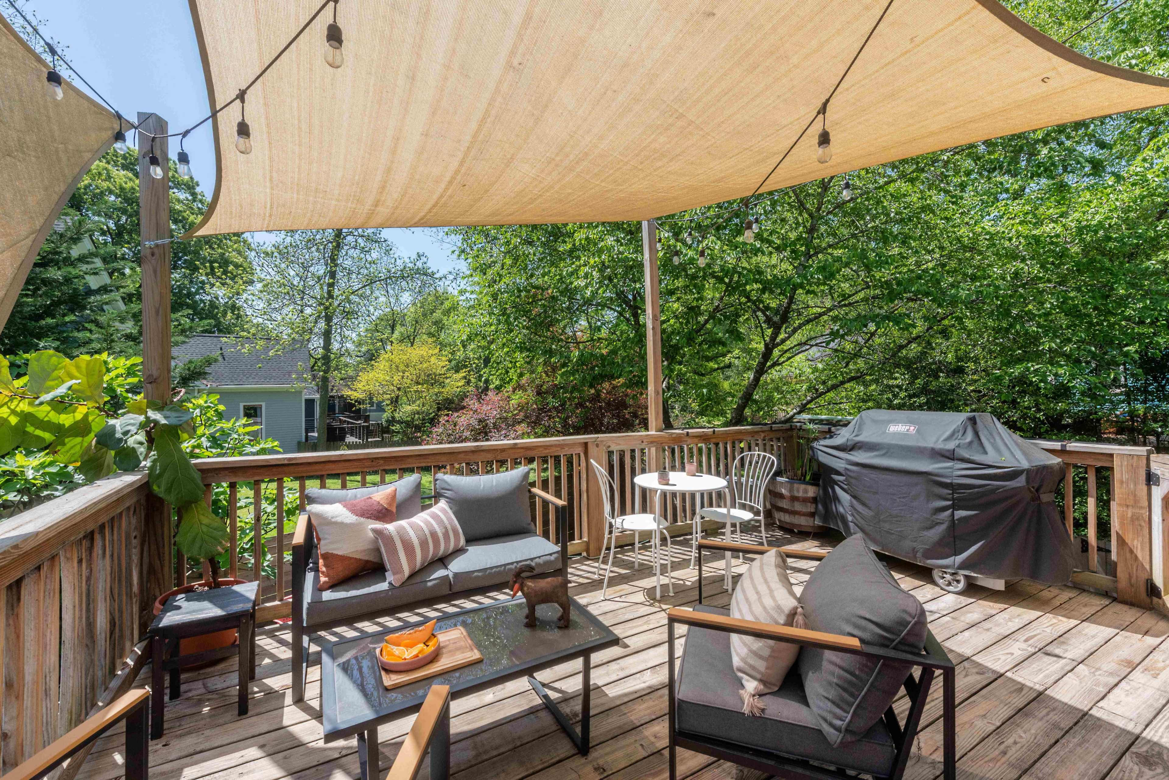 A wooden deck features seating areas with cushions, a small table, and a grill, all shaded by large sail-like canopies surrounded by lush greenery.