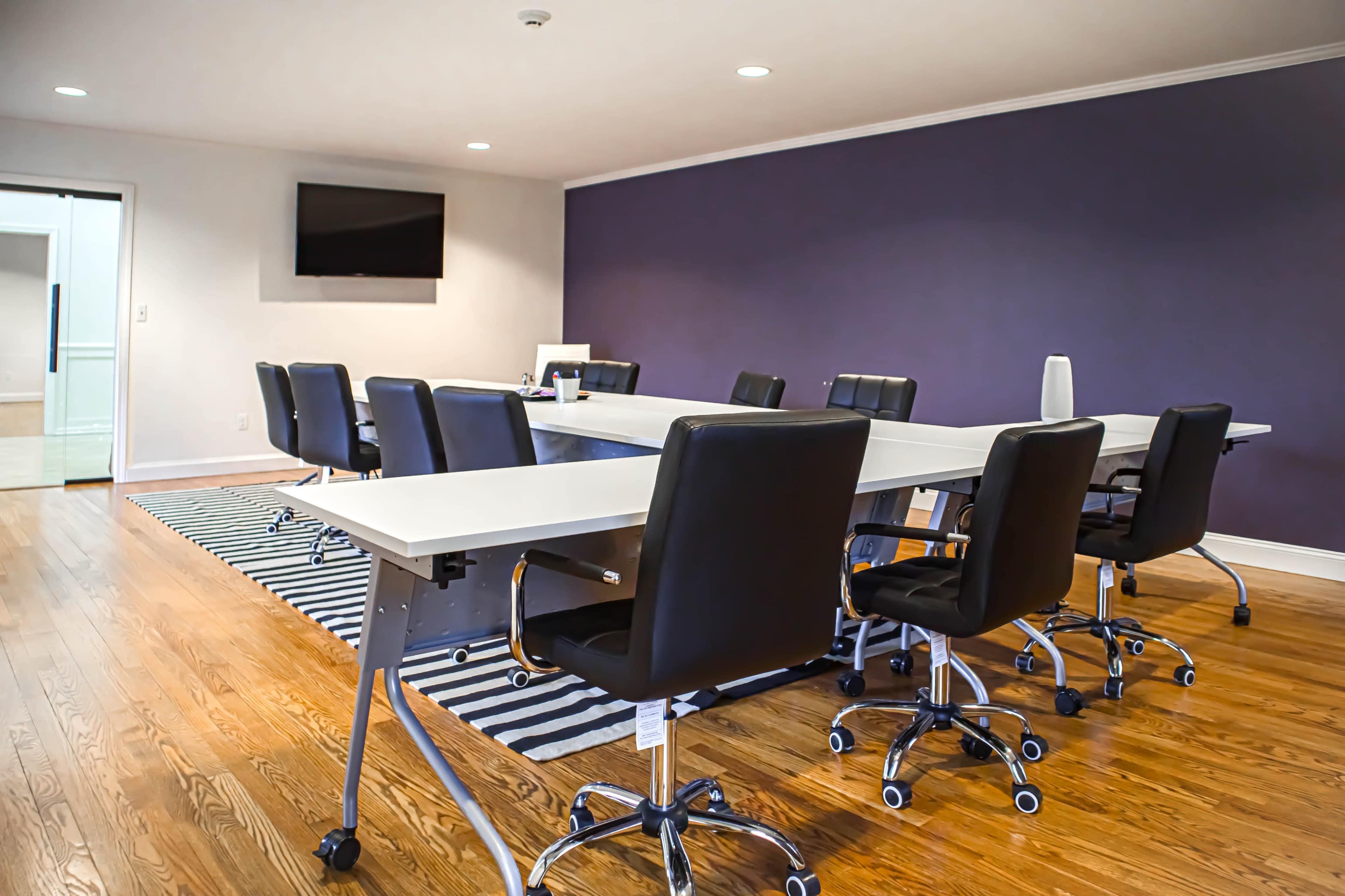 A modern conference room featuring a central long table surrounded by black rolling chairs, with a television mounted on the wall and a decorative rug beneath the table.