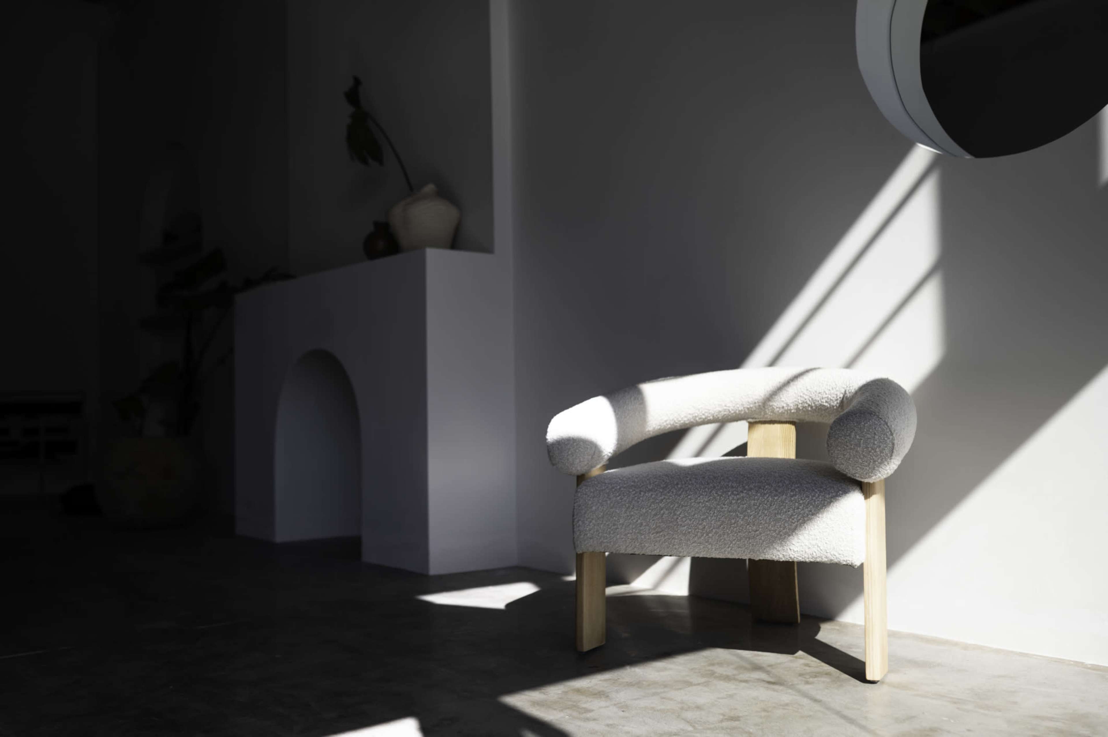 A light-colored, upholstered chair with rounded arms is positioned in a room with minimal decor and shadows cast by a window.
