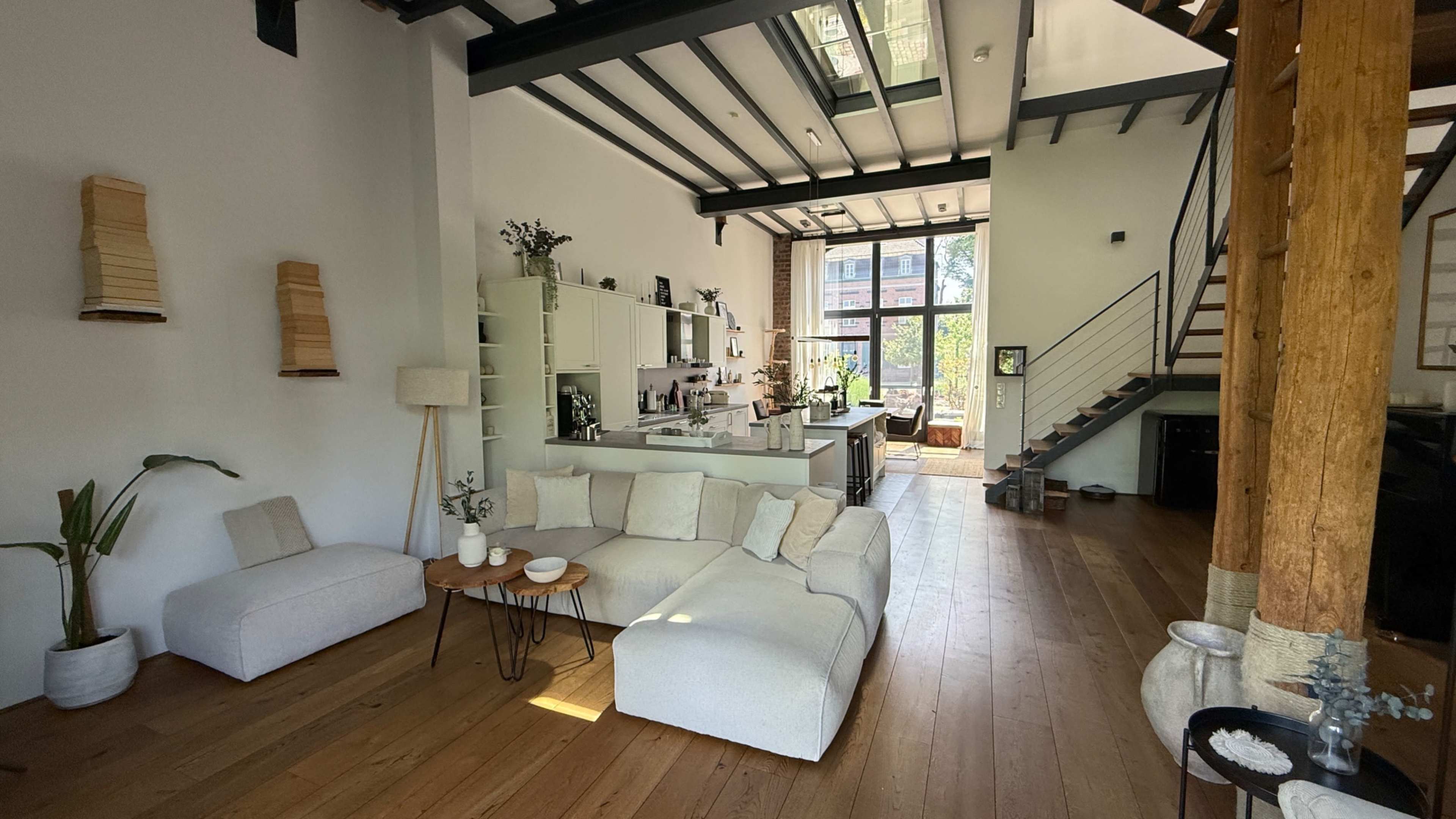 The image shows a modern interior living space with a sectional sofa, wooden flooring, and a kitchen area featuring large windows that allow natural light to enter.