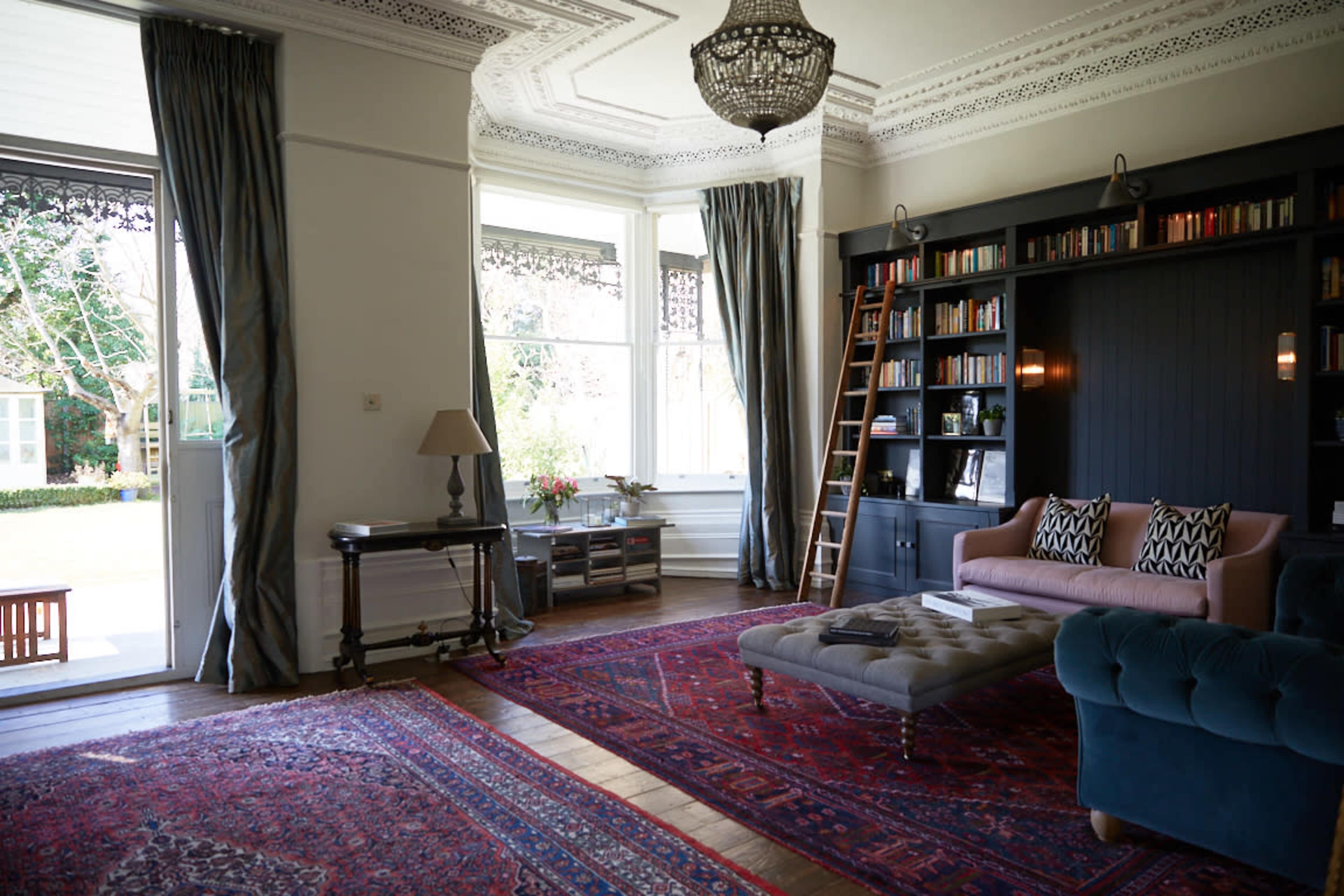 The scene depicts a stylish living room featuring a large bookshelf, a sofa, a coffee table, and a decorative area rug, with large windows opening to a garden view.