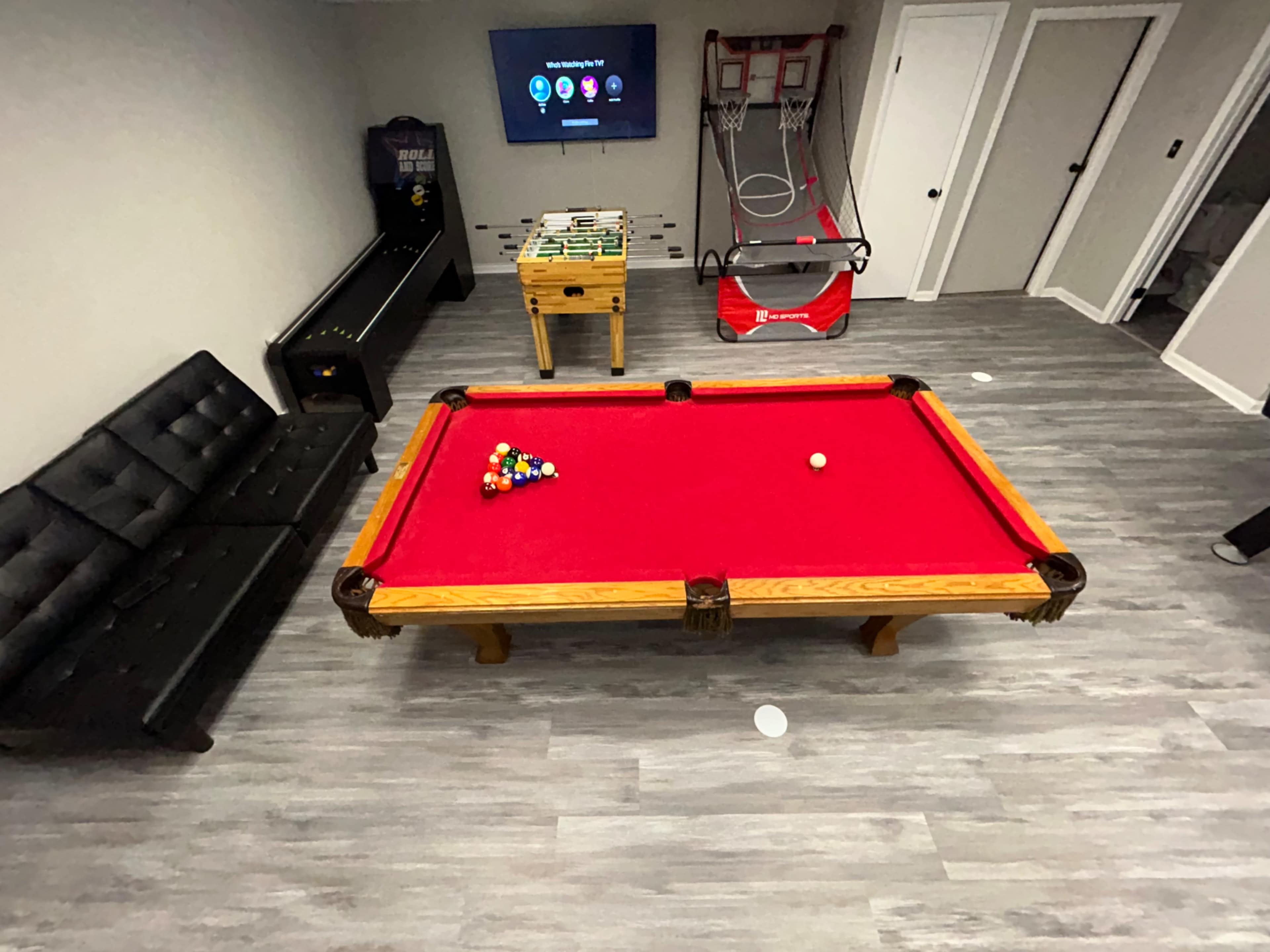 The image shows a game room with a red pool table at the center, a black couch on the left, a TV mounted on the wall, and a basketball arcade game in the background.