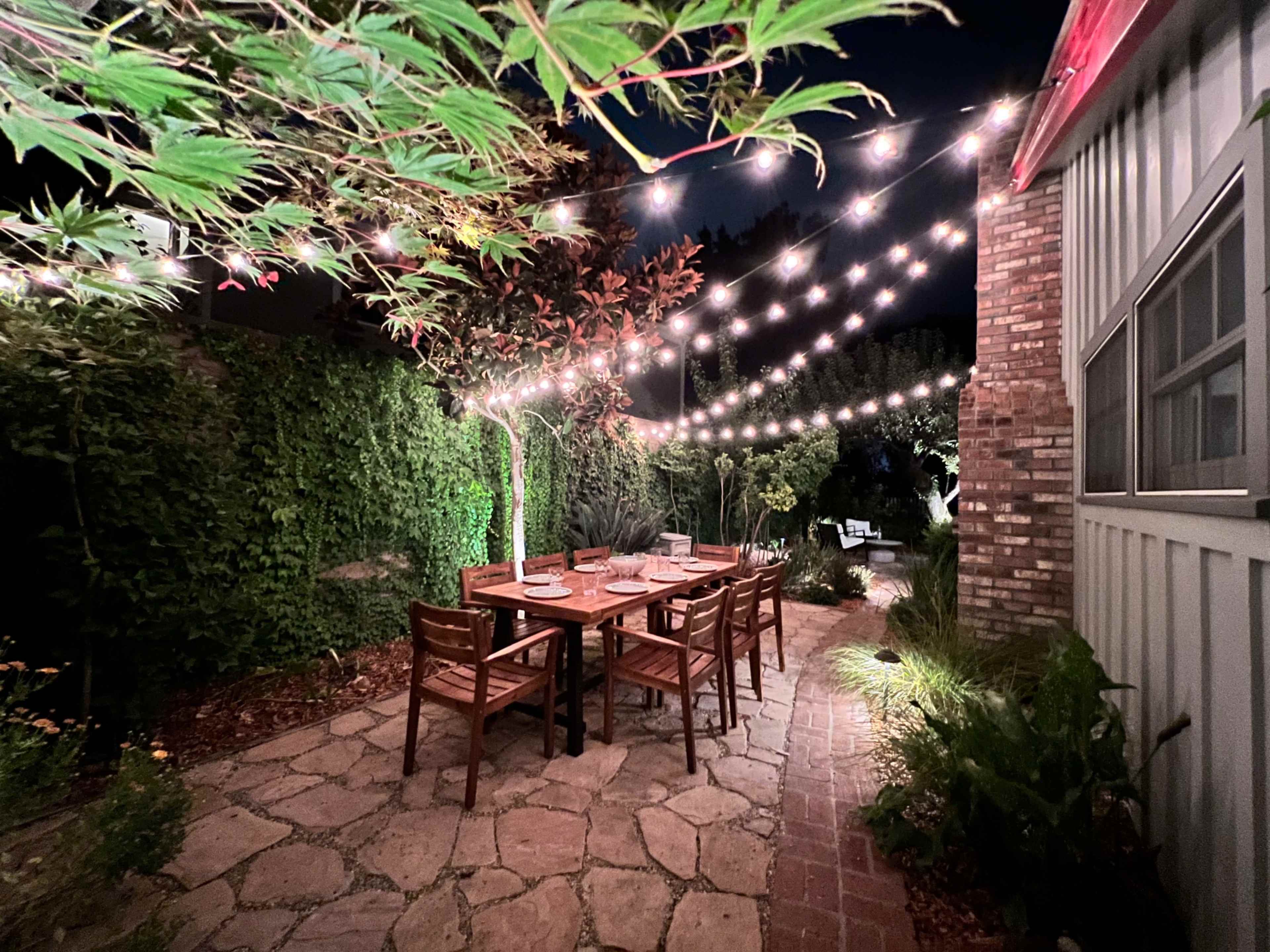 A wooden dining table surrounded by chairs is illuminated by string lights in a garden at night.