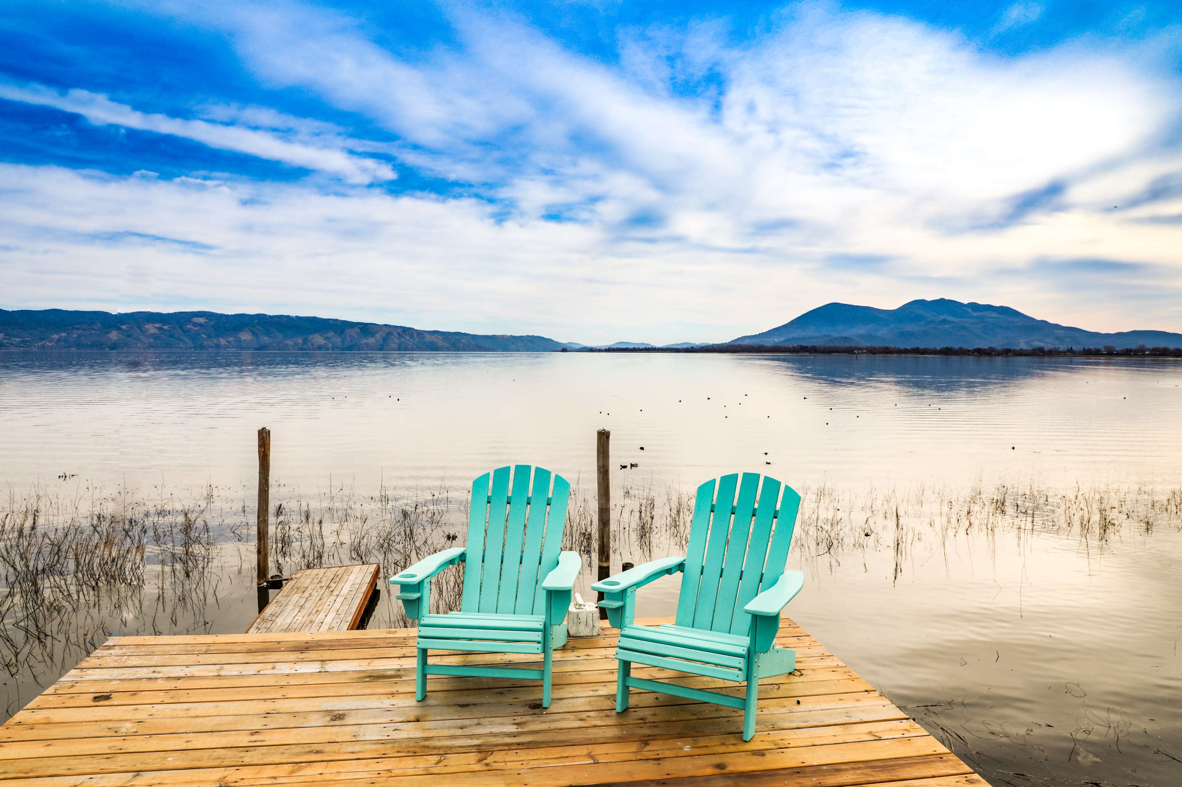 Two turquoise Adirondack chairs are positioned on a wooden dock overlooking a calm lake with mountains in the background.