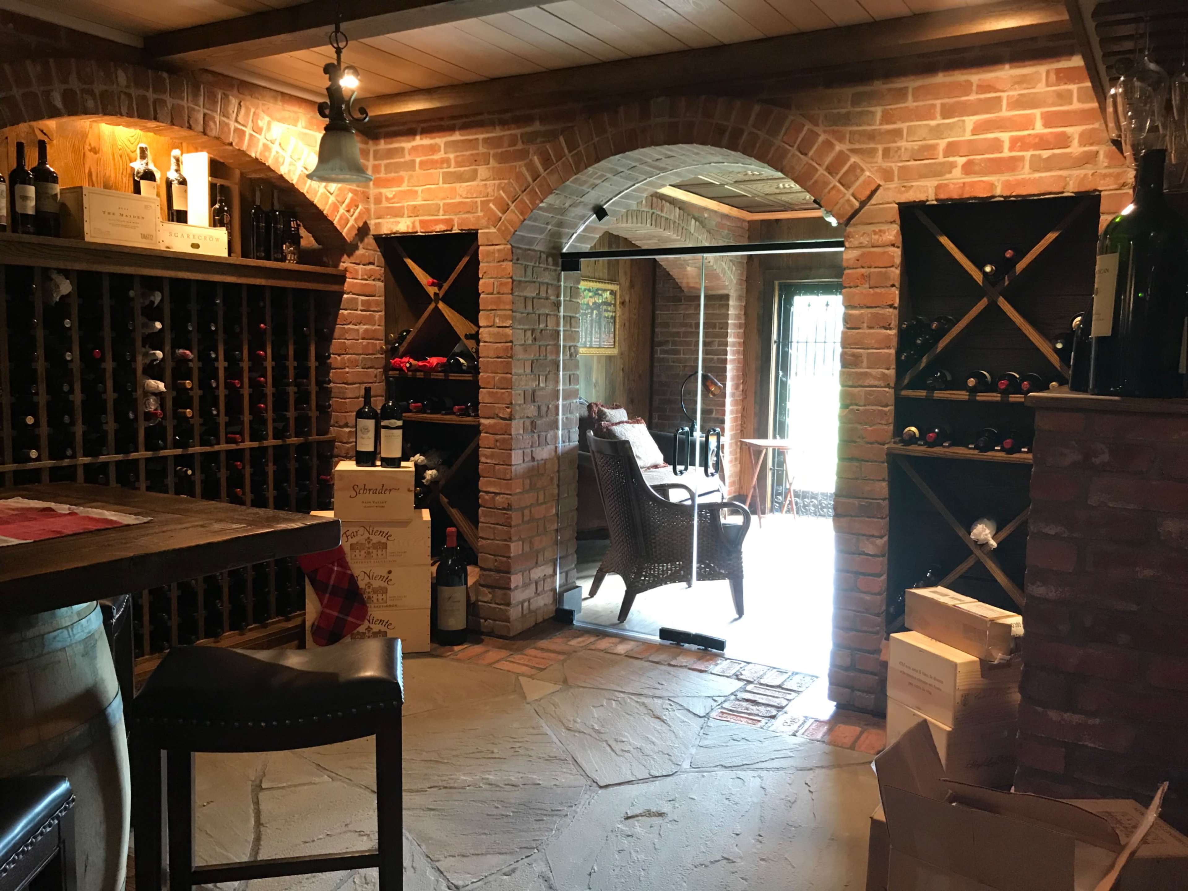 A wine cellar with brick walls, wooden shelves filled with wine bottles, and a seating area visible through an arched doorway.