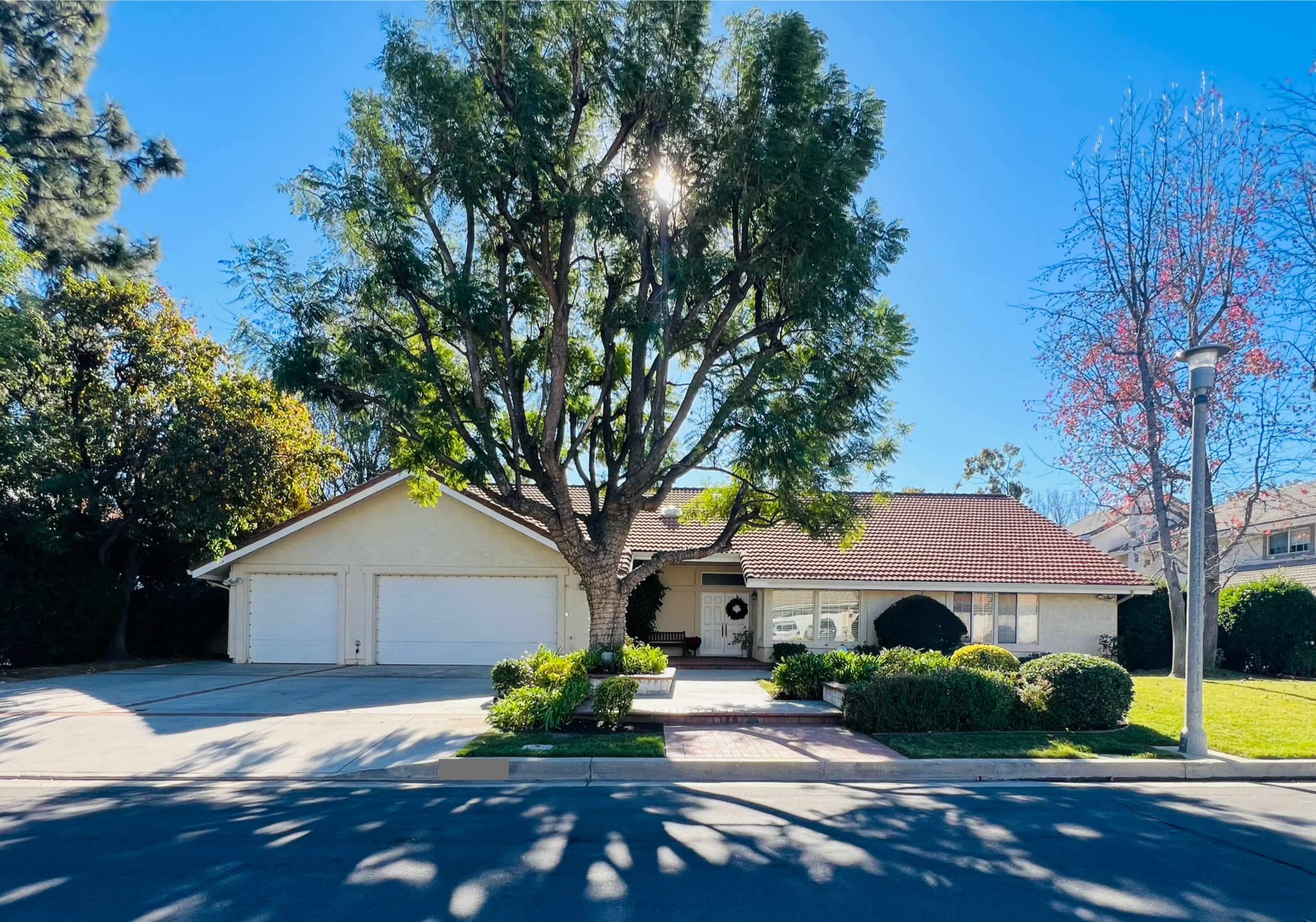 A single-story house with a red-tiled roof and two garage doors is surrounded by neatly trimmed shrubs and a large tree in the front yard.