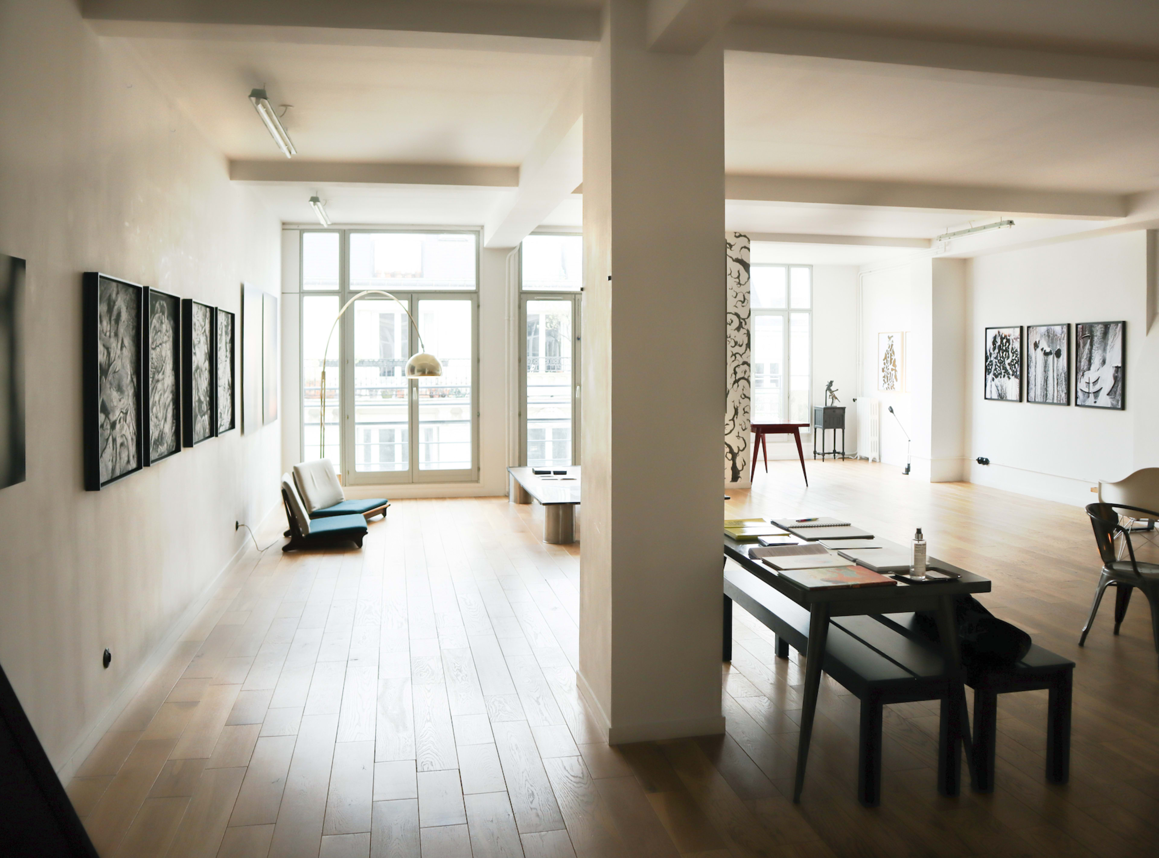 The image shows a modern, spacious art studio with wooden flooring, featuring several artworks displayed on the walls and a table with art supplies in the center.