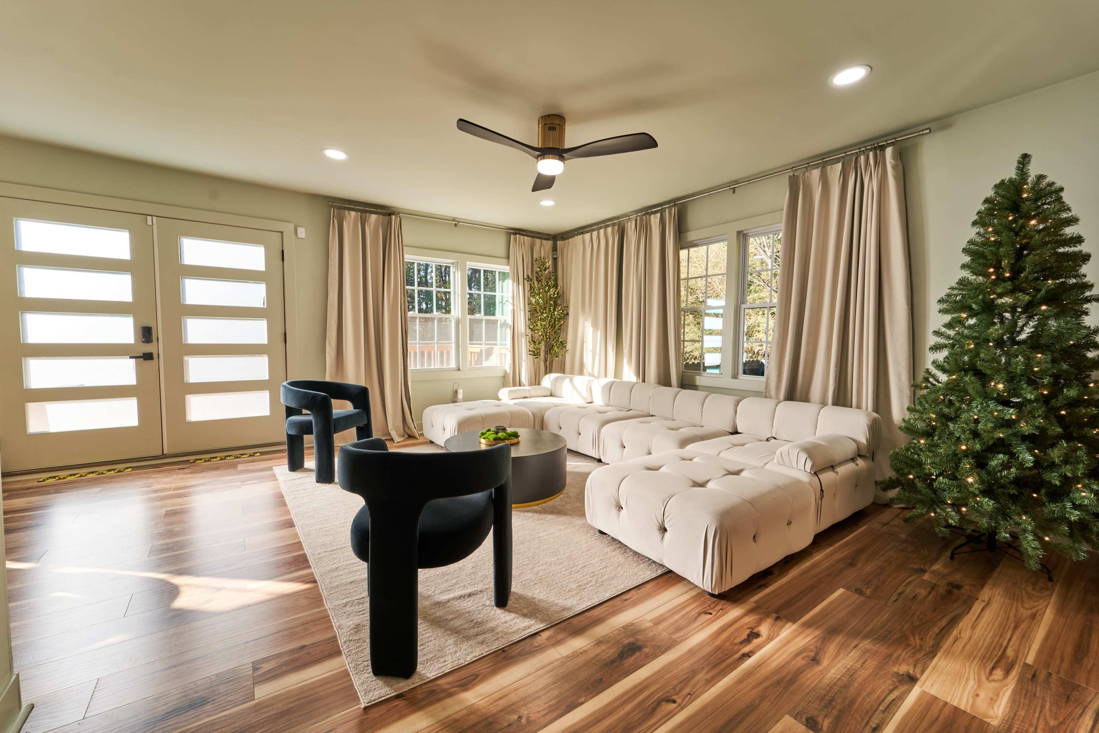 A modern living room features a light-colored sofa, black accent chairs, a coffee table, and a Christmas tree beside large windows.