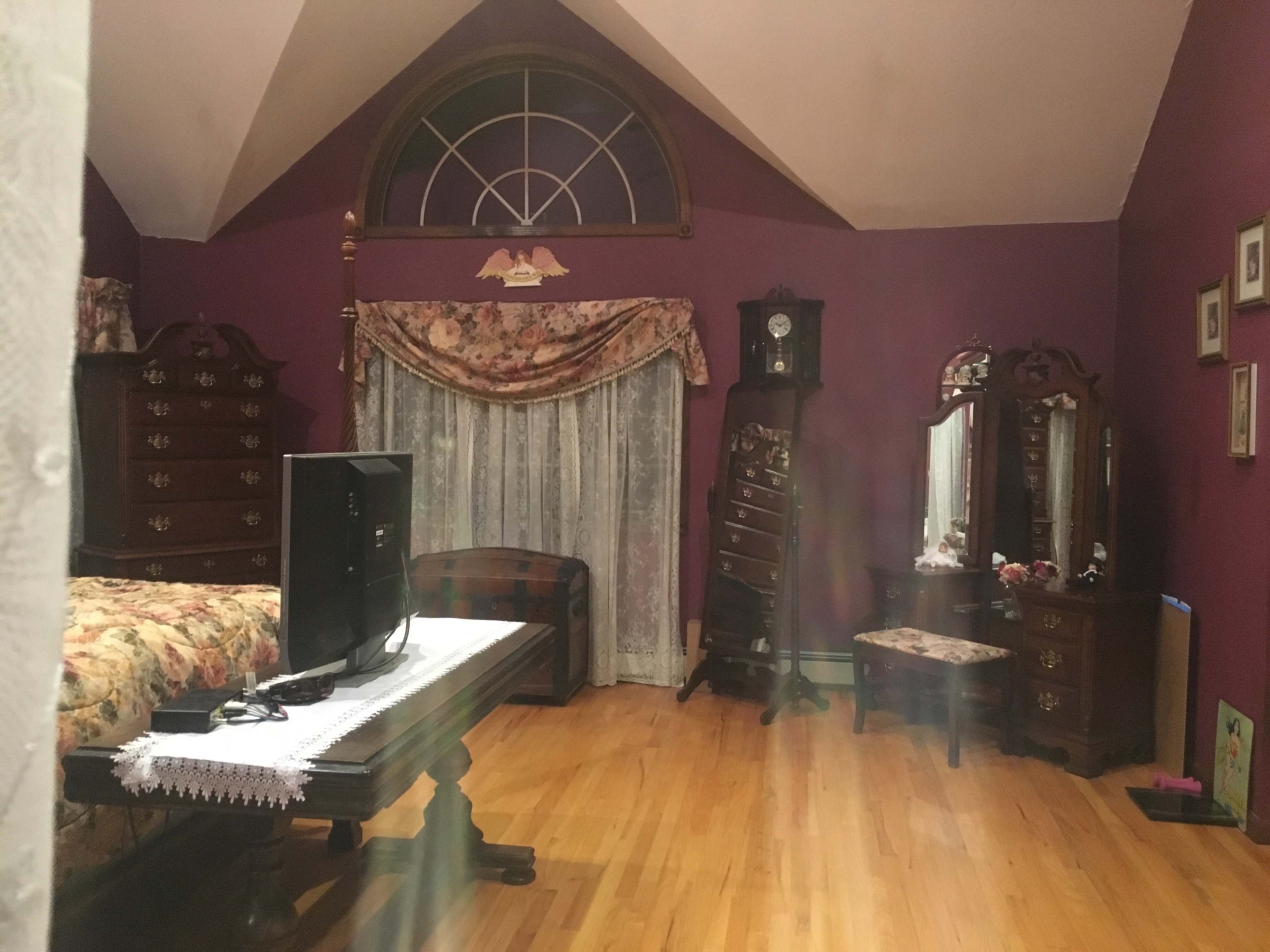 The room features a bed with a floral comforter, a television on a wooden stand, and various antique furniture pieces against purple walls.