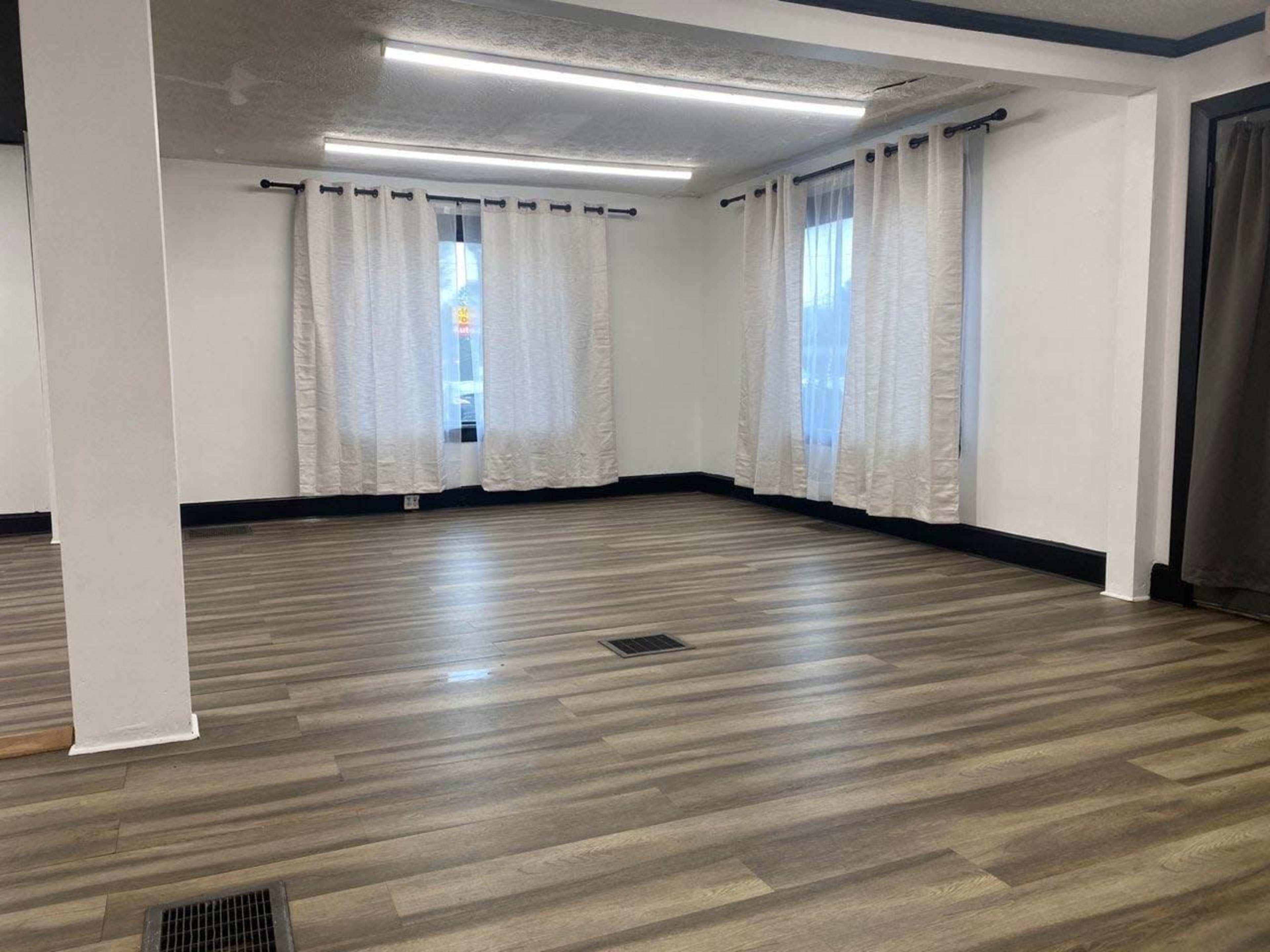 The image shows a spacious room with wooden flooring, large windows dressed in white curtains, and a minimalist design.