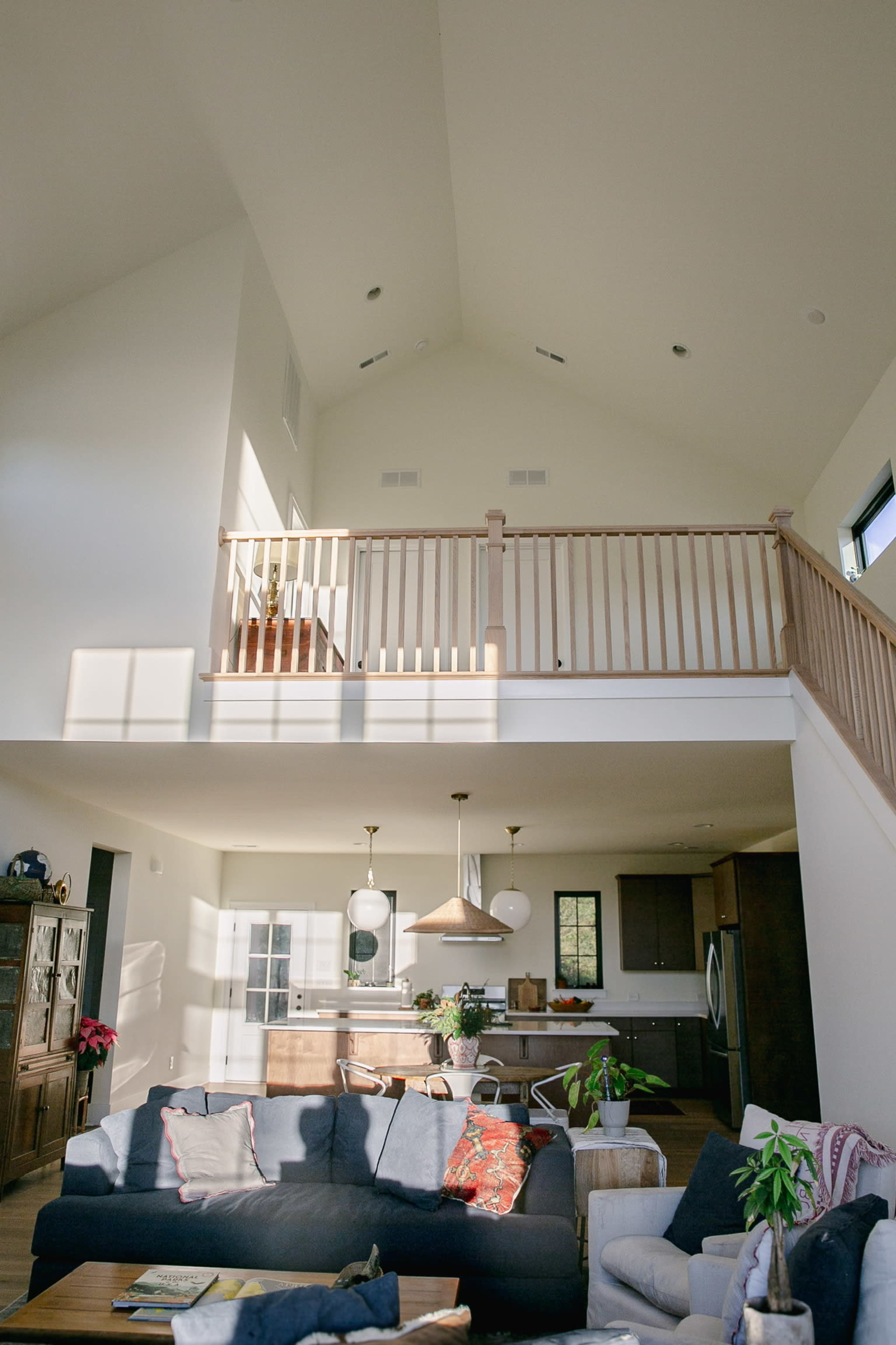The image shows a spacious living area with high ceilings, featuring a balcony, a cozy sofa, and a kitchen area in the background.