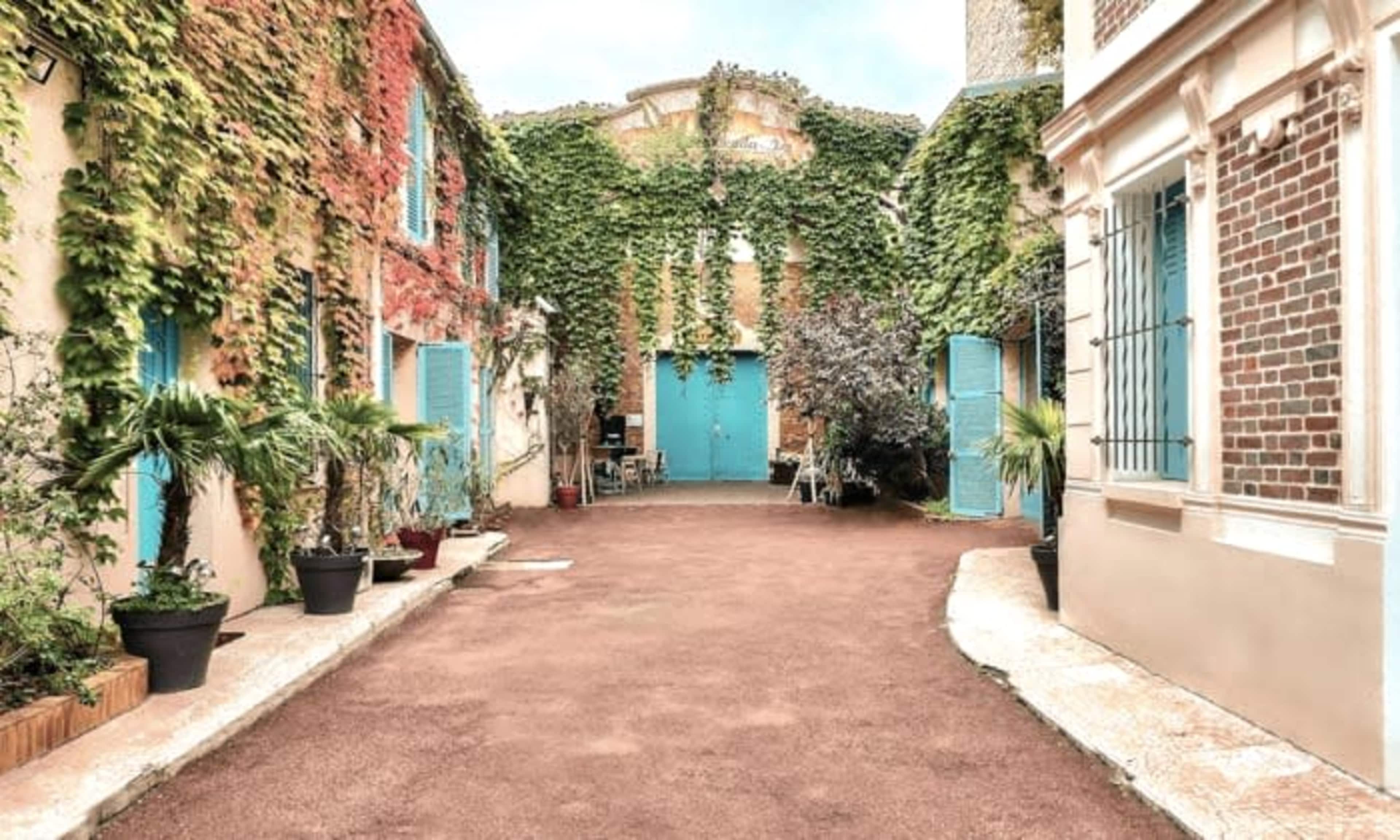 The scene shows a narrow pathway lined with buildings covered in vines, featuring blue doors and potted plants on either side.