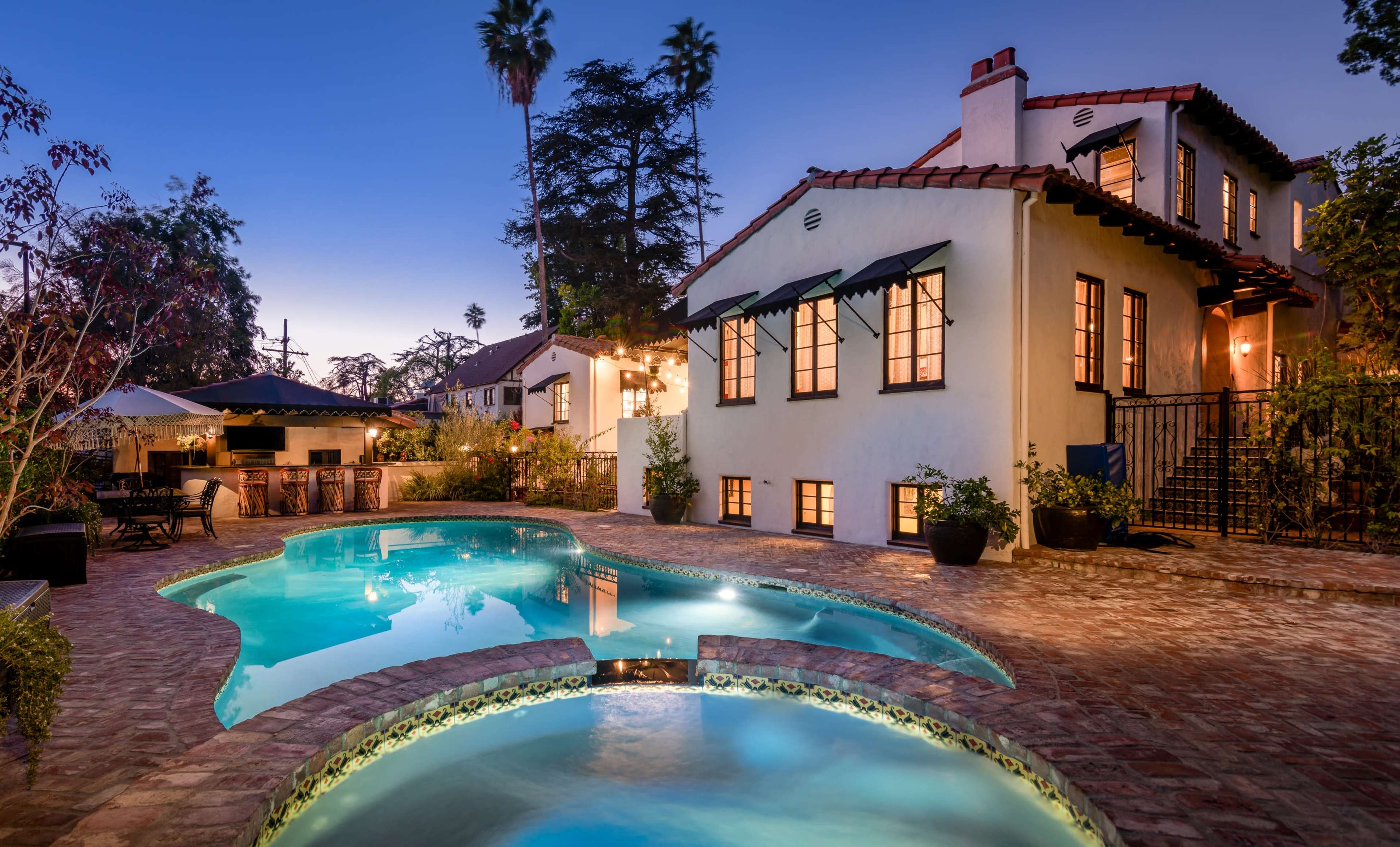The image shows a well-lit home with an inviting pool and spa area, surrounded by landscaping and palm trees in a twilight setting.