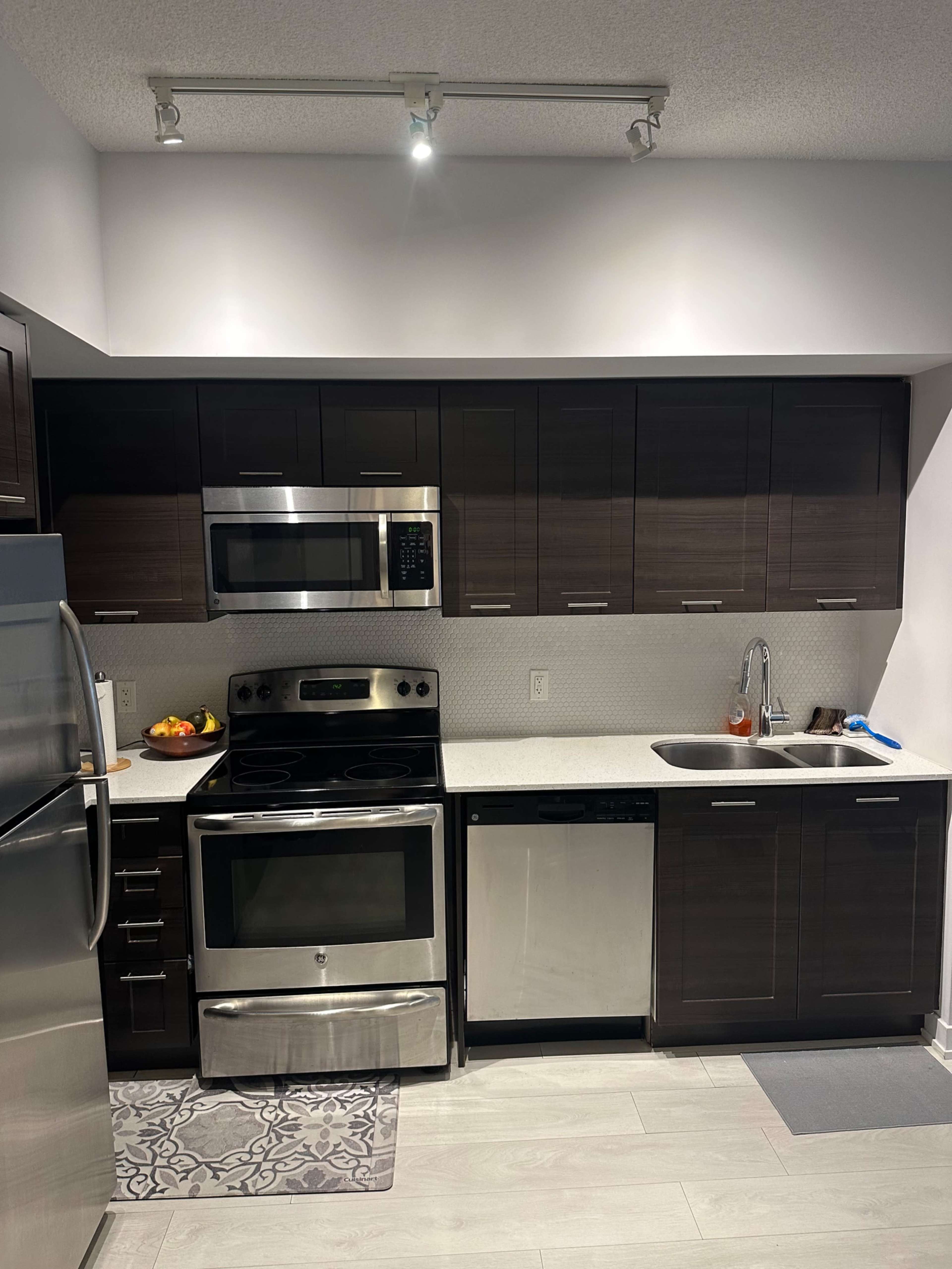 The image shows a modern kitchen with dark wood cabinetry, stainless steel appliances, and a countertop with a sink and fruit bowl.
