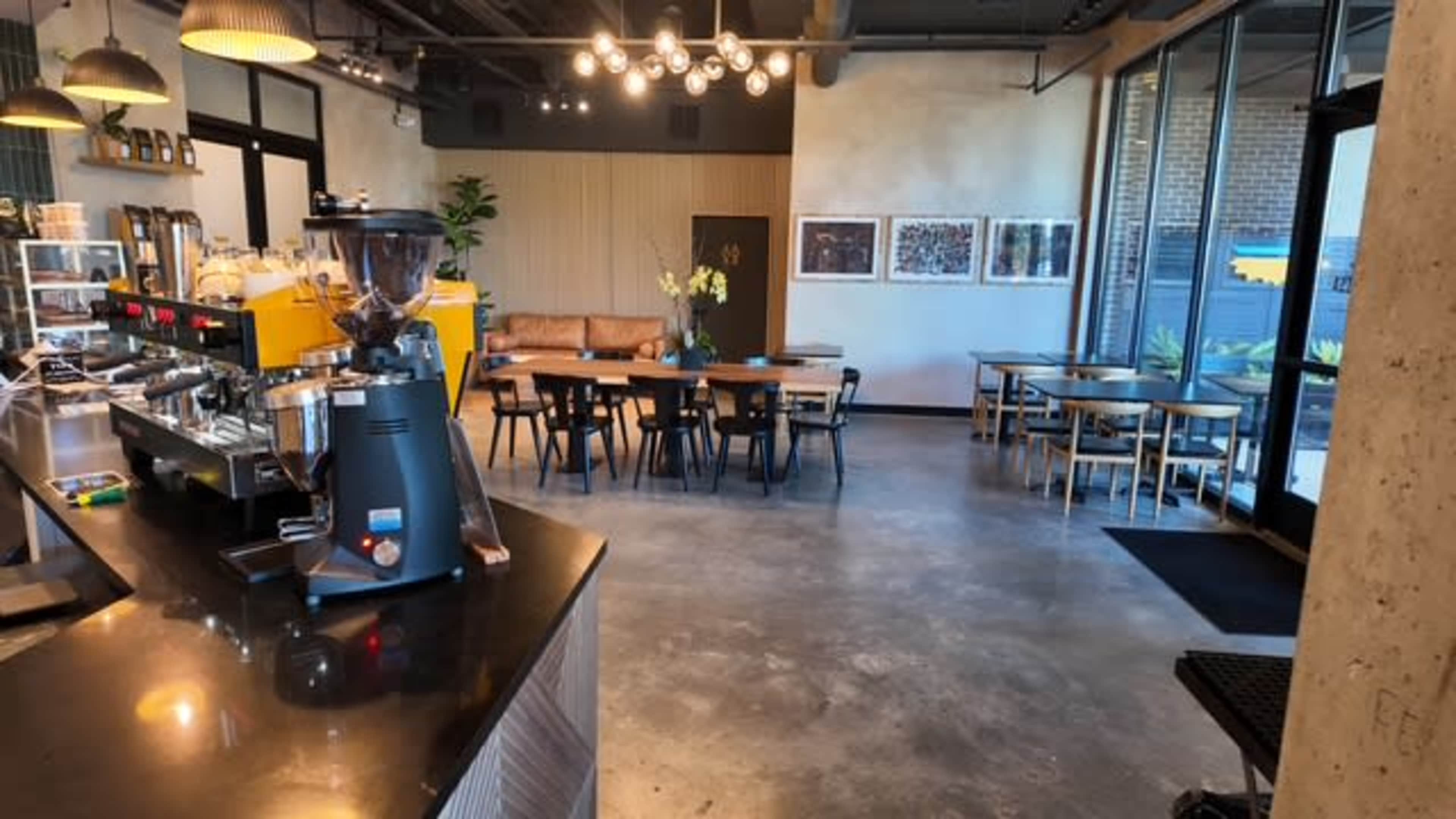 The image shows a modern café interior with a coffee machine on the counter, seating arrangements including tables and a couch, and large windows allowing natural light to enter the space.