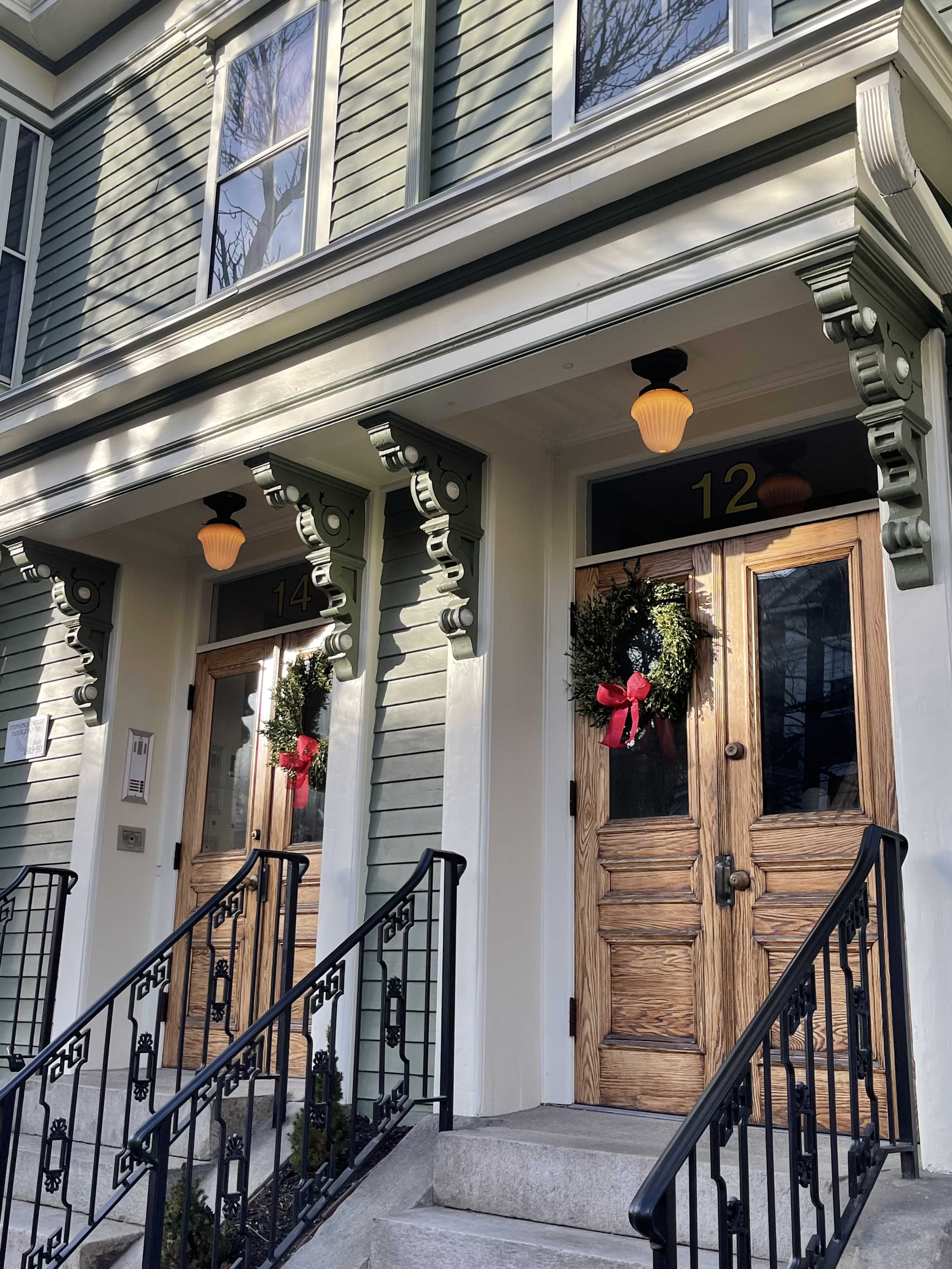 Two front doors of a green wooden house, each adorned with wreaths and flanked by decorative light fixtures.