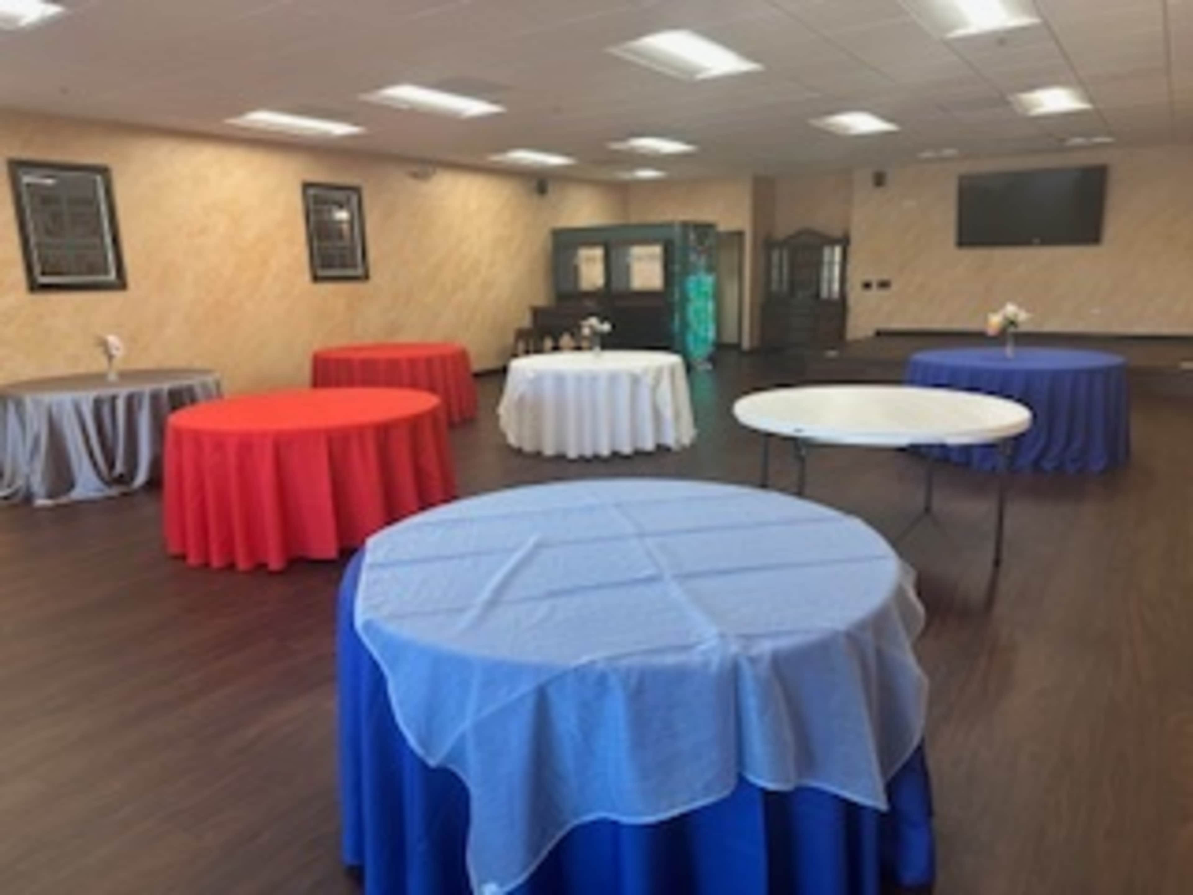 The image shows a room with several round tables covered in blue, red, white, and gray tablecloths, arranged neatly on a wooden floor.