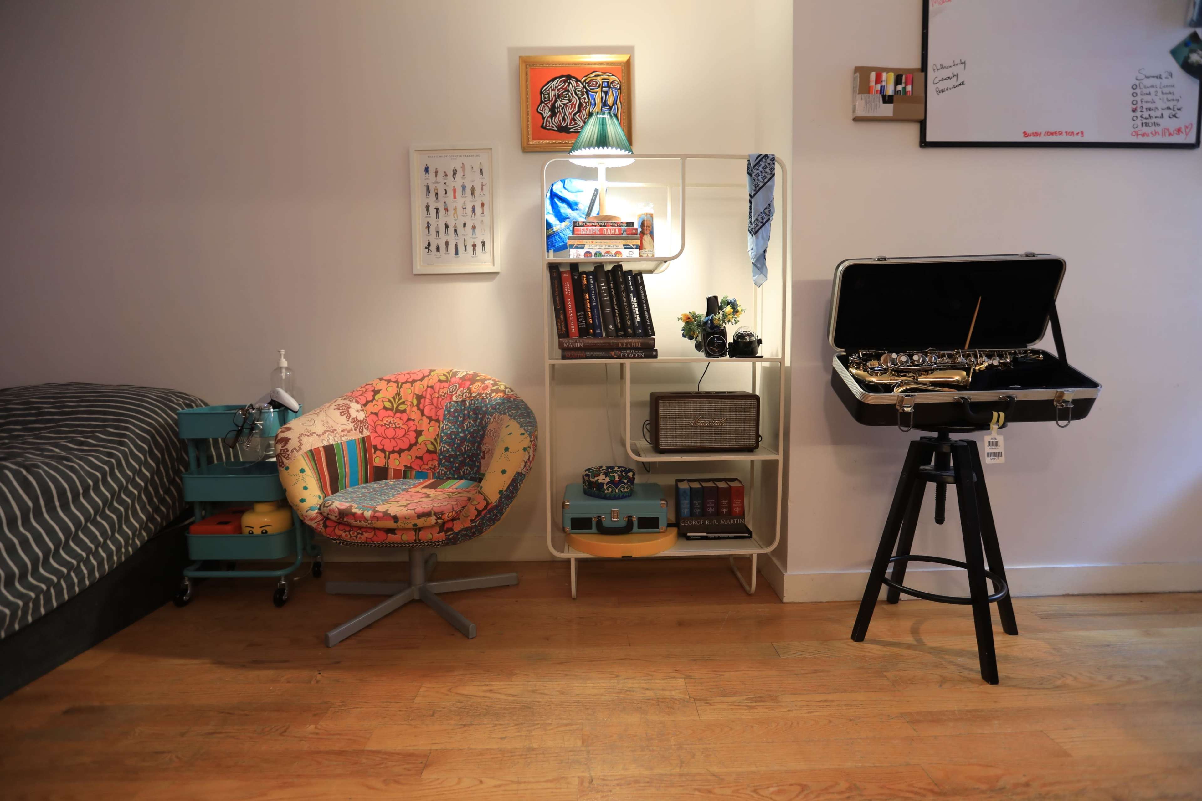 A small room featuring a colorful chair, a bookshelf filled with various items, a mirror, and a suitcase-style stand beside a bed with striped bedding.