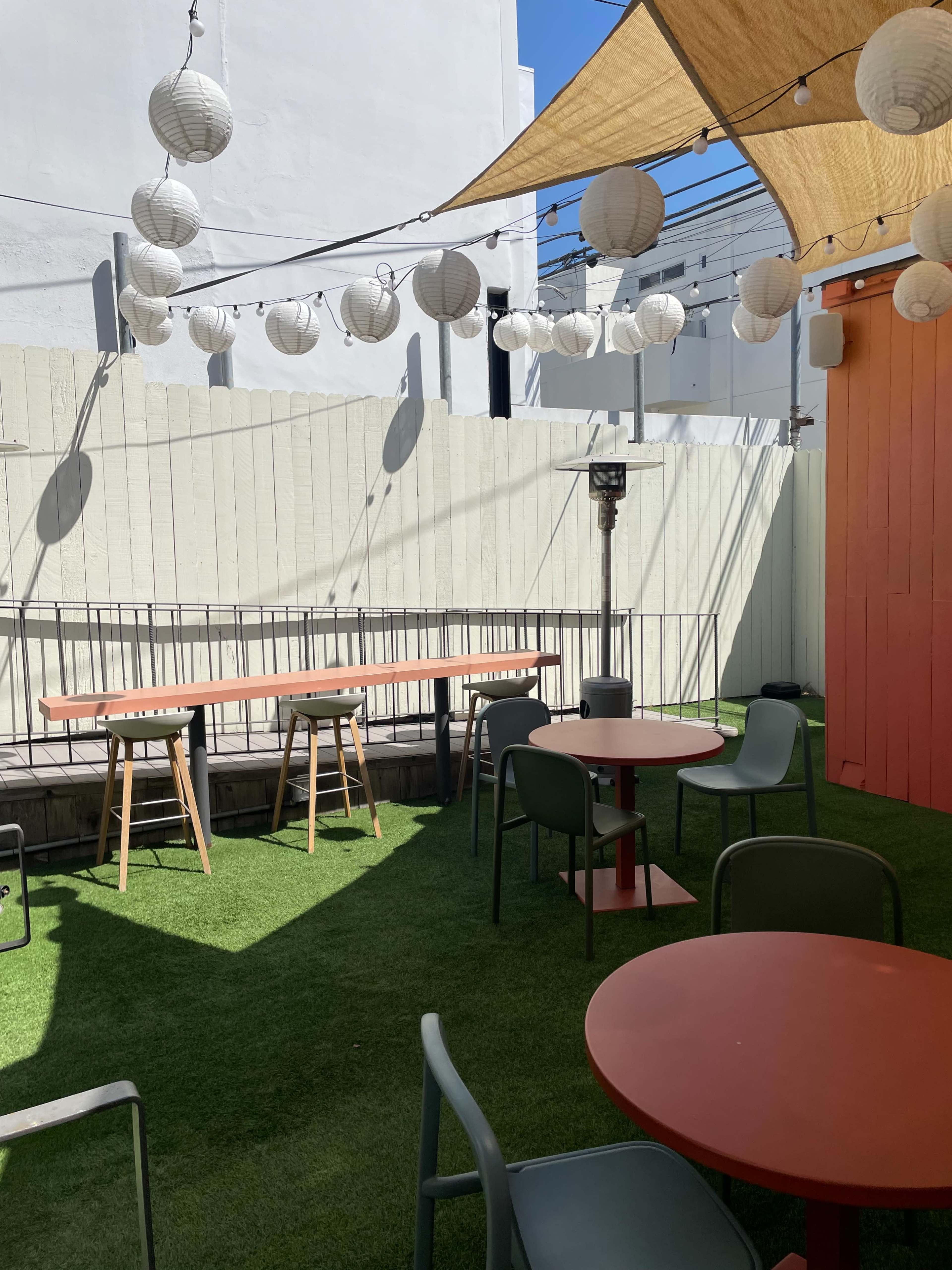 An outdoor patio area features green artificial turf, scattered tables, and lanterns hanging above.