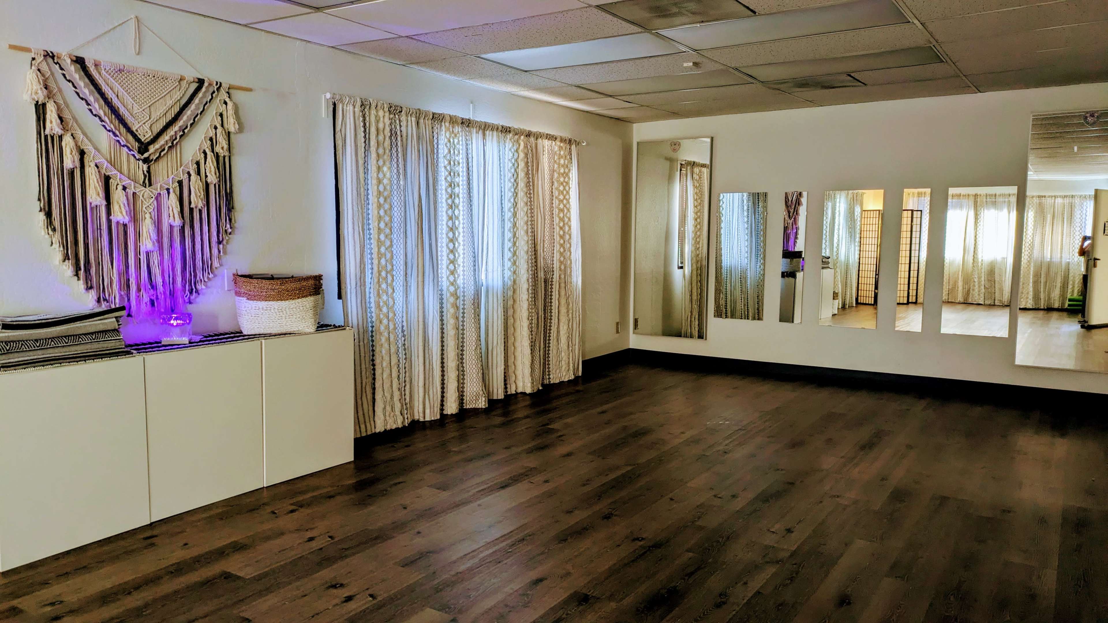The image shows a spacious, well-lit room with wooden flooring, large windows covered with sheer curtains, and a decorative wall hanging beside a cabinet and mirrors on the opposite wall.