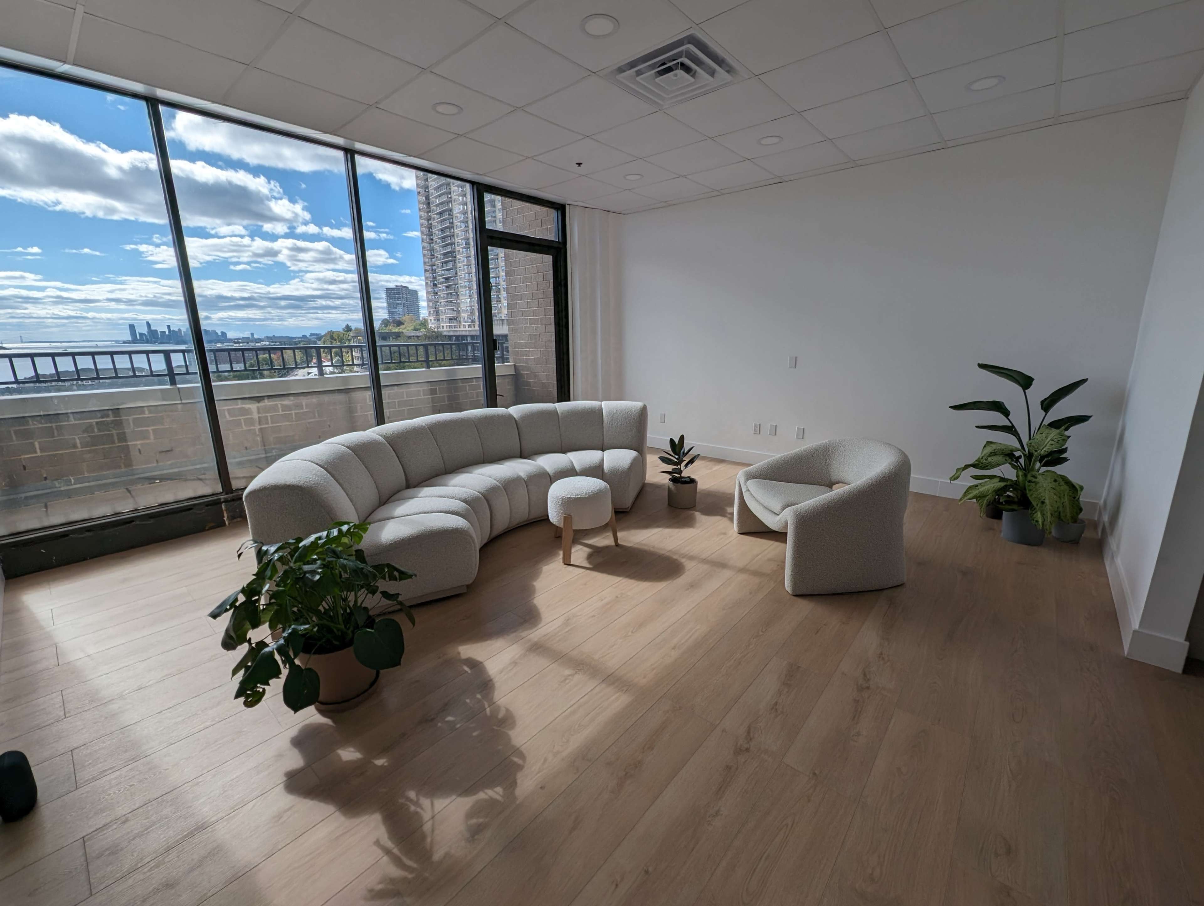 A modern living room features a curved sofa, a small round table, and a single armchair, all surrounded by potted plants and large windows displaying a city skyline.