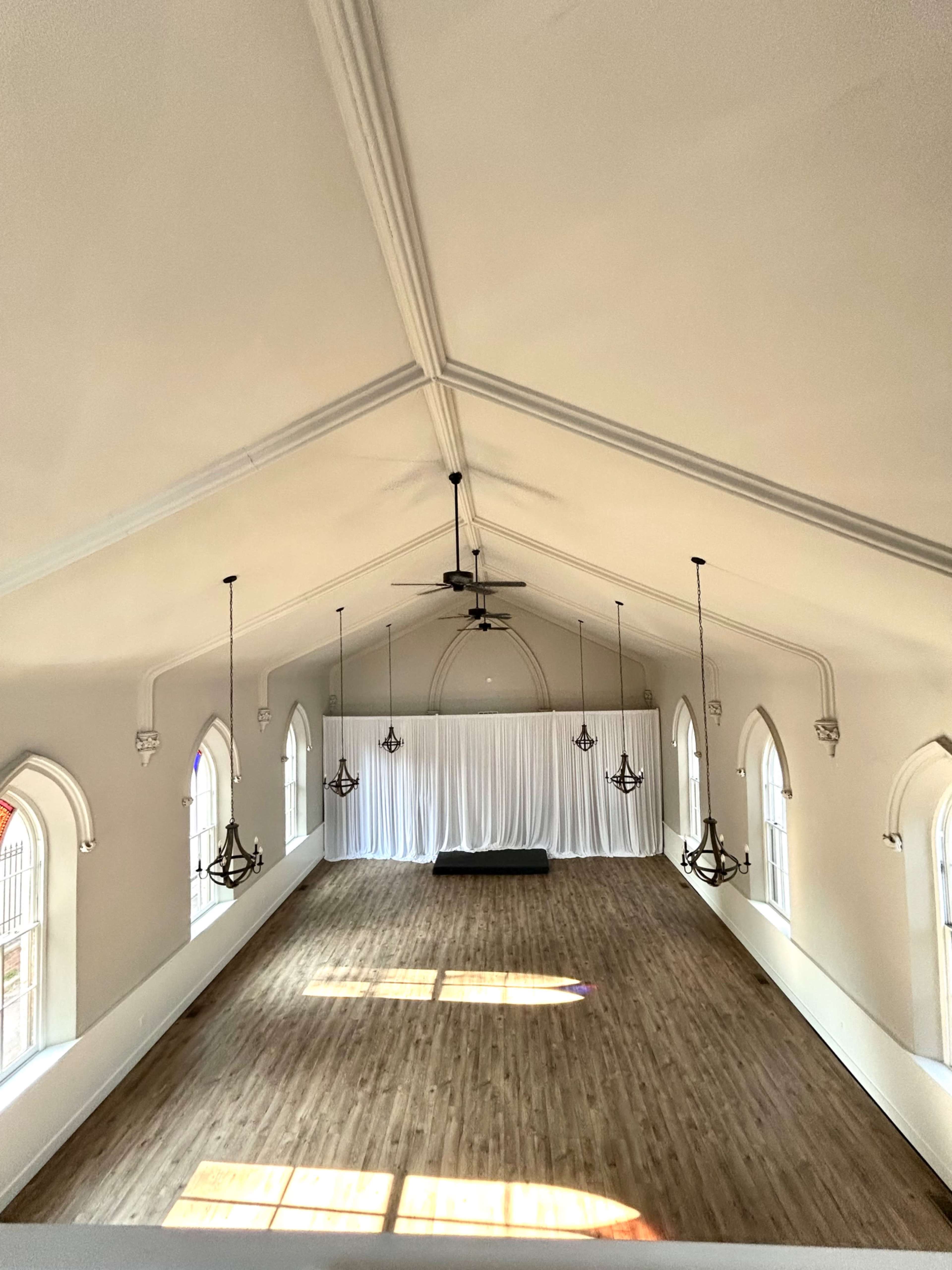The image shows a spacious indoor hall with a vaulted ceiling, decorative chandeliers, large windows, and a hardwood floor.