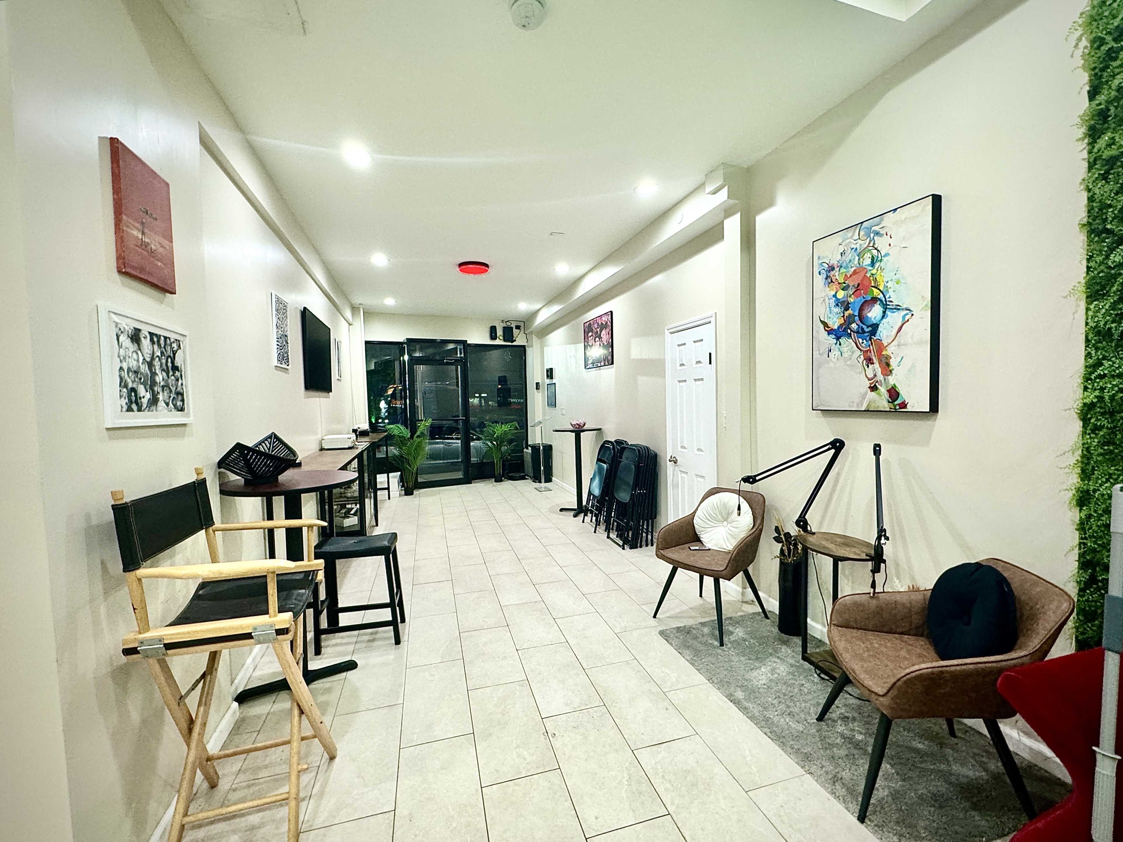 The image shows a spacious, well-lit interior with modern furnishings, including chairs and tables, along one side and artwork on the walls.