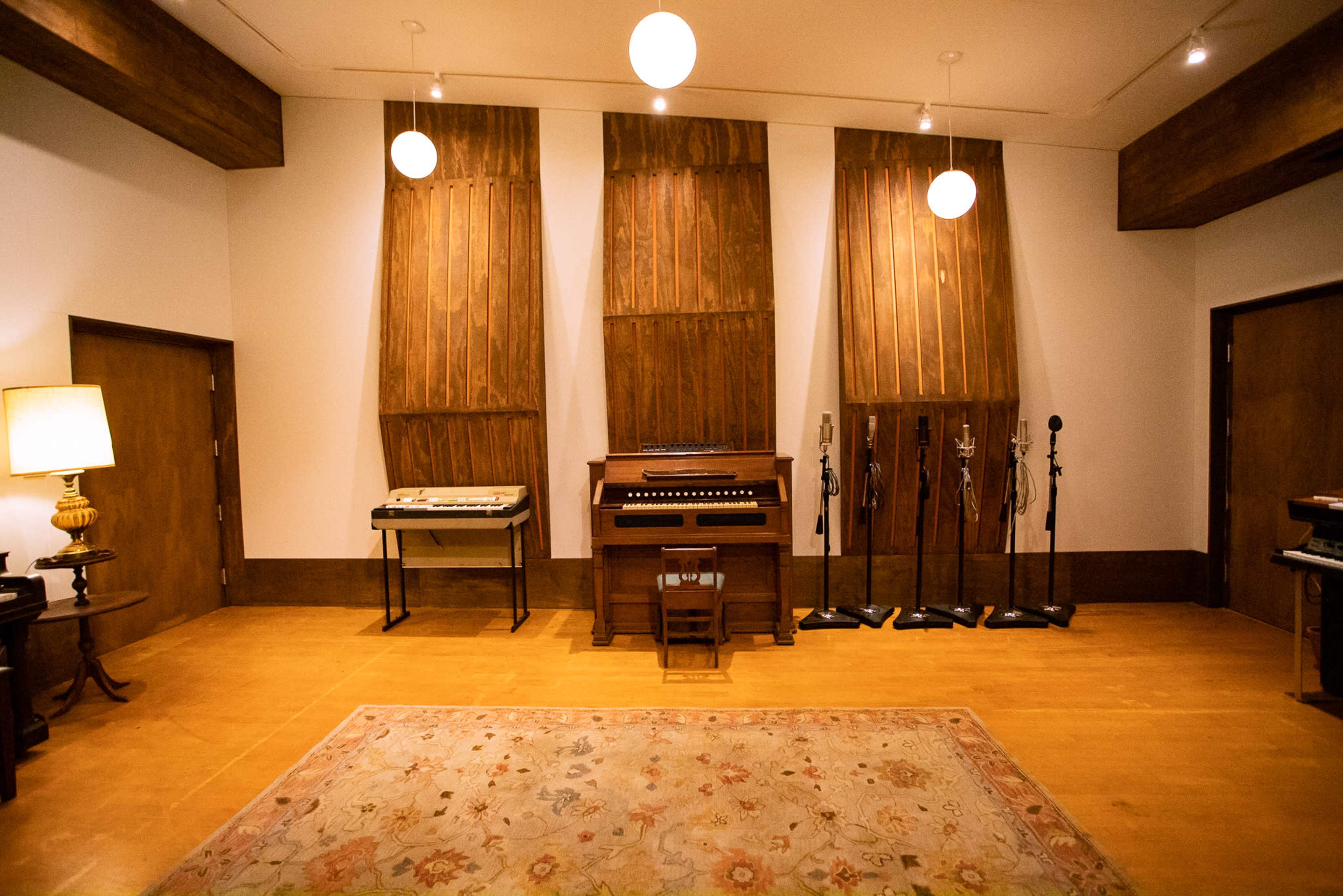 A spacious music recording room featuring wooden wall panels, a vintage organ, and several microphones on stands.