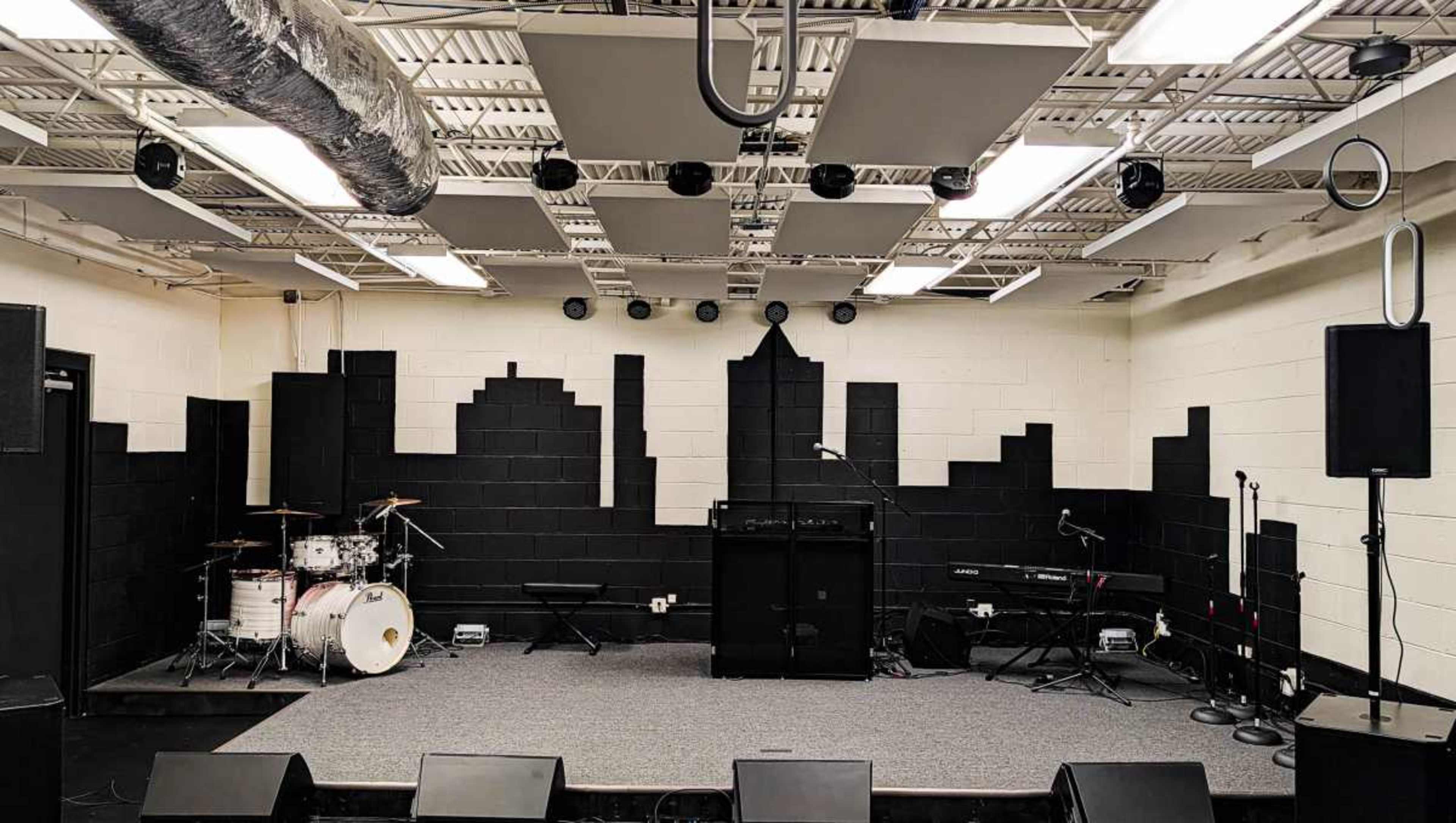 The image shows a rehearsal space featuring a stage with a drum set, an amplifier, and two microphones in front of a black silhouette of a city skyline on the back wall.