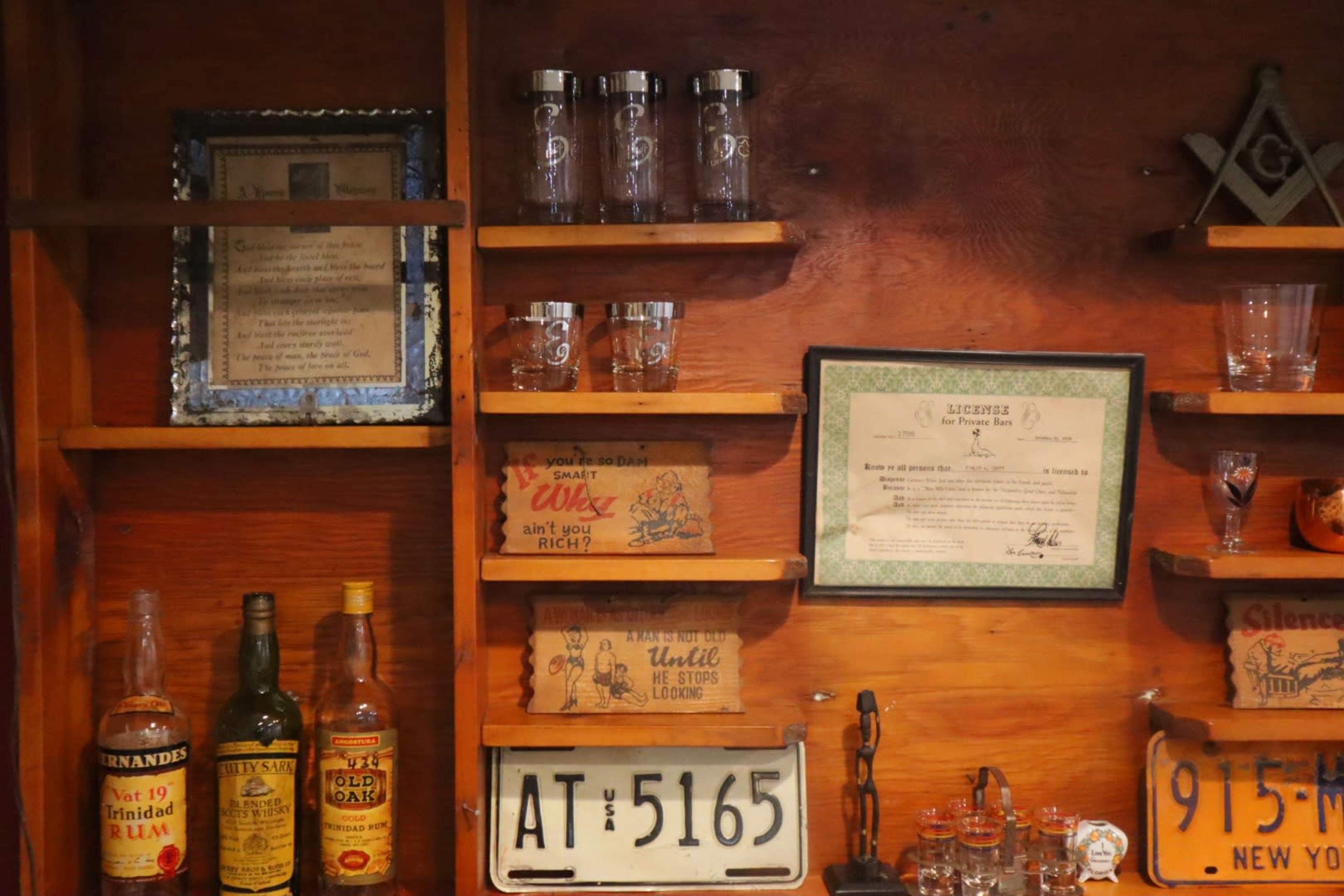 A wooden shelf displaying various bottles of liquor, glassware, and decorative signs alongside a framed certificate and a vintage license plate.
