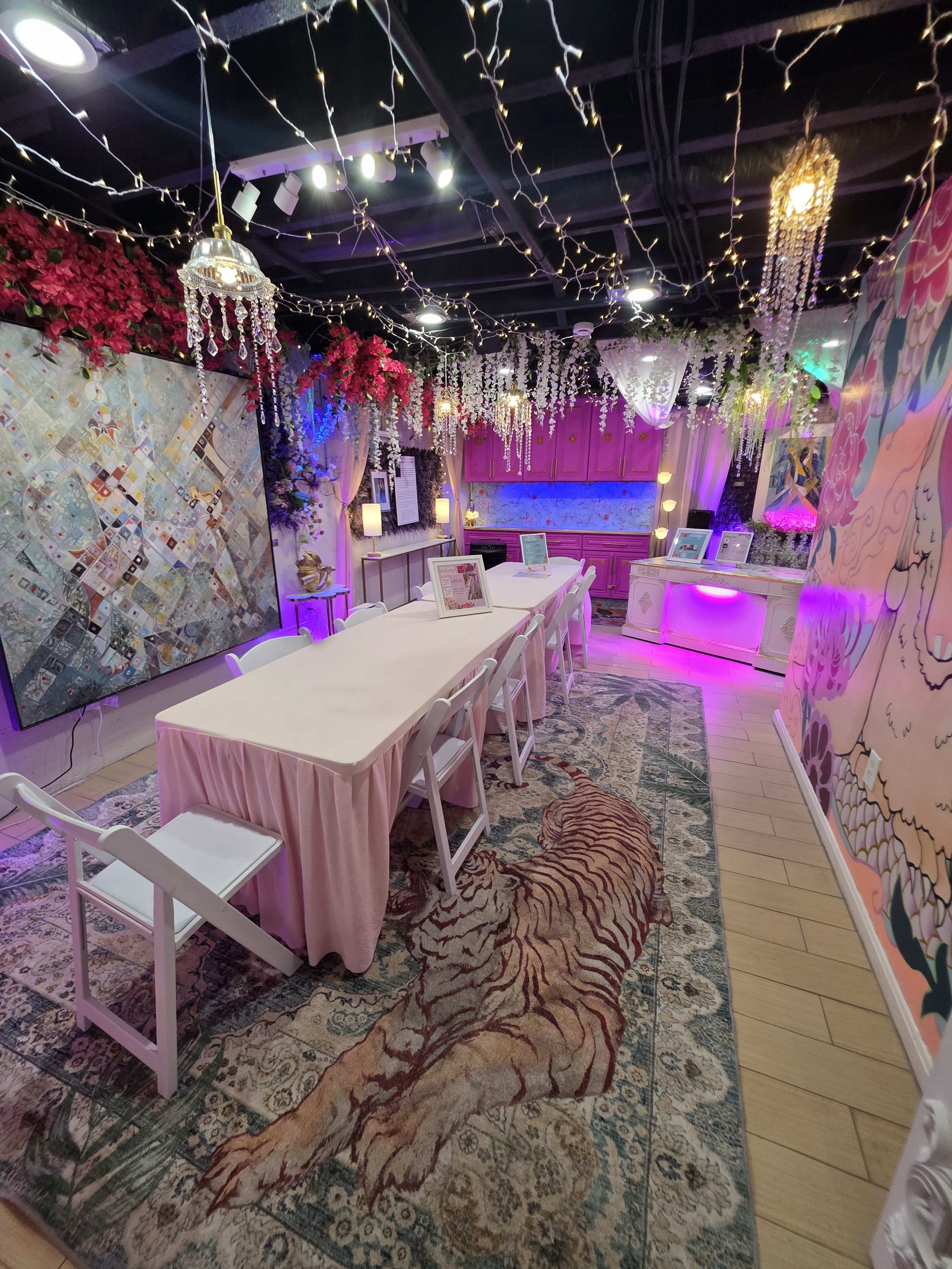 The image shows a brightly decorated indoor space with a long table covered by a tablecloth, surrounded by chairs, and adorned with hanging lights and floral decorations.