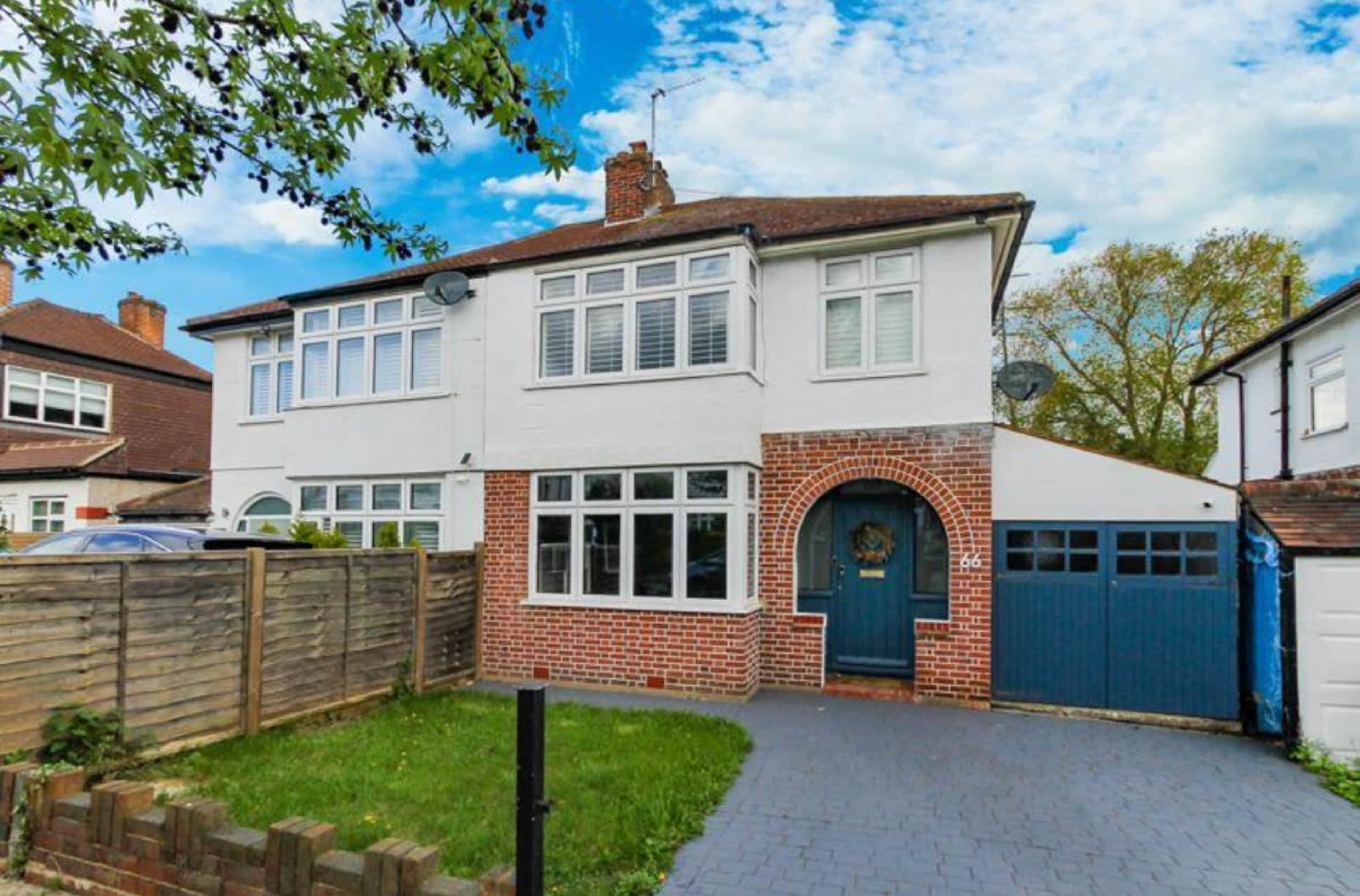 The image shows a two-story, semi-detached house with a brick and white exterior, a blue garage door, and a front garden featuring a grassy area and a wooden fence.