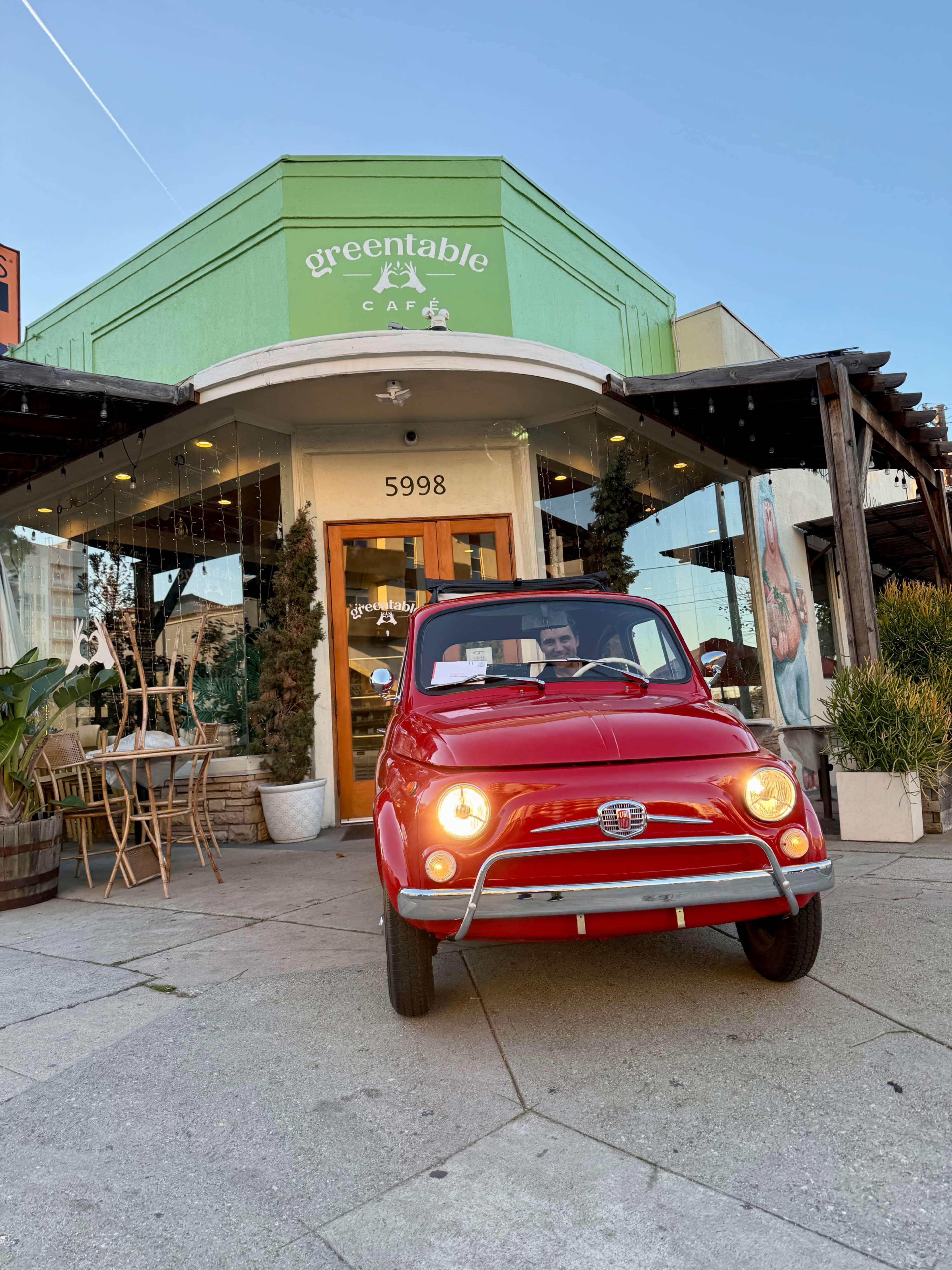 A red vintage car is parked in front of Greentable Café, which features outdoor seating and a welcoming entrance.