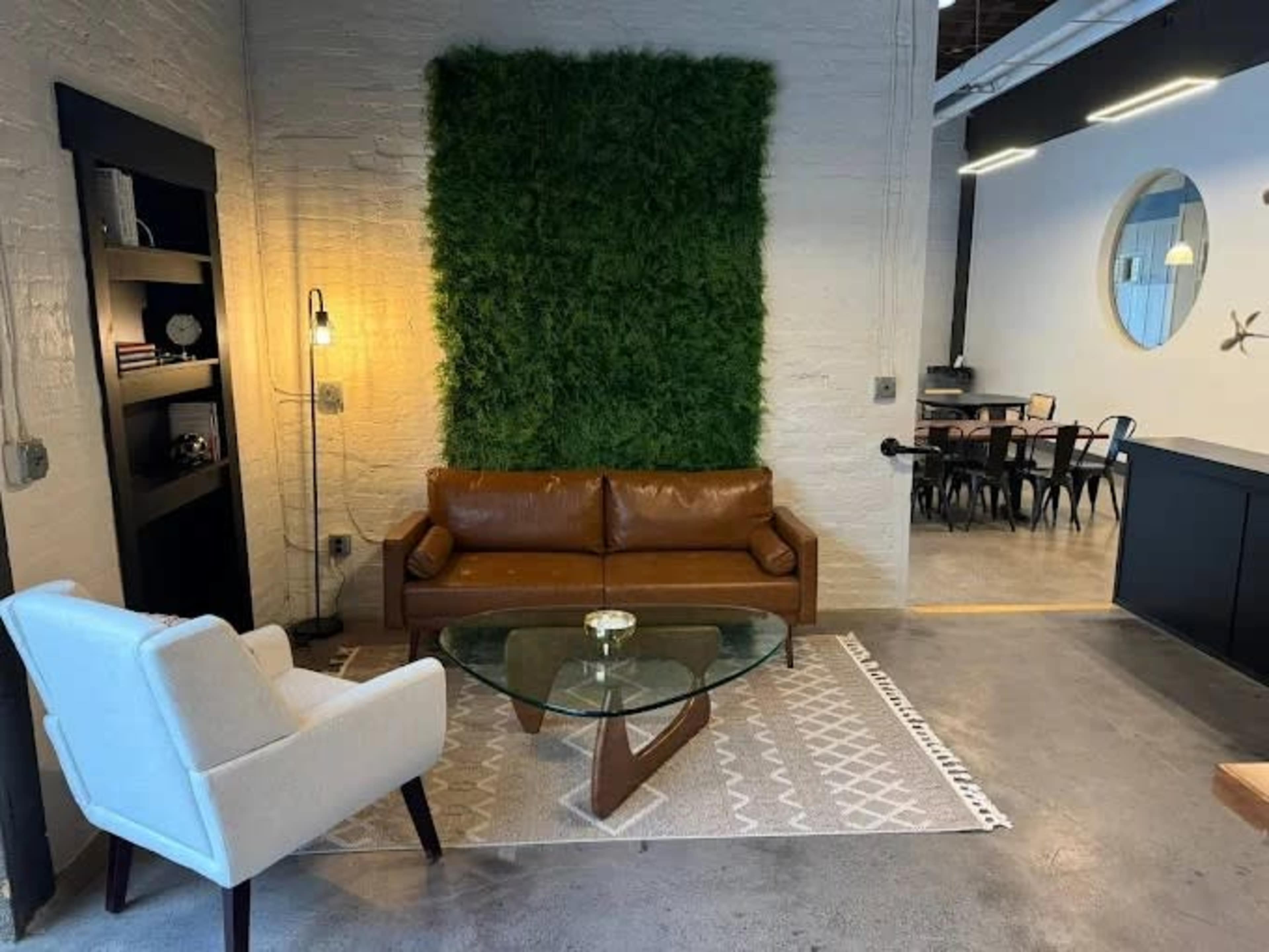 The image shows a modern living space featuring a brown leather sofa, a glass coffee table on a patterned rug, and a green wall accent made of artificial plants.
