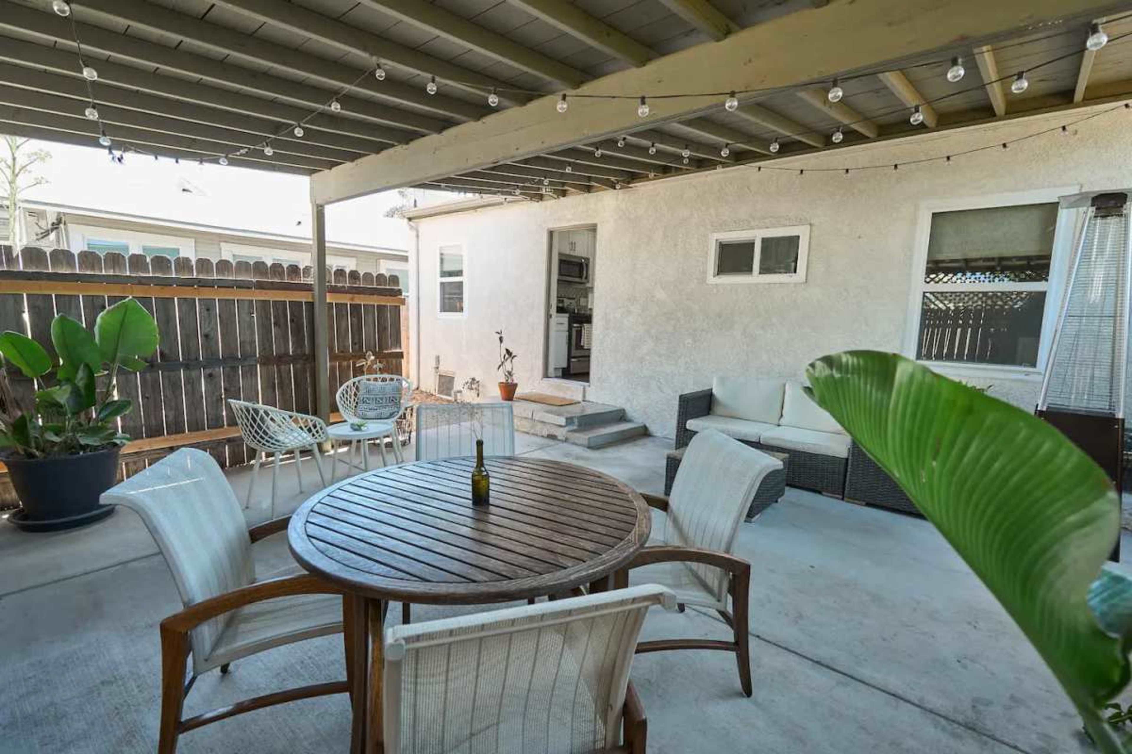 The image shows a covered outdoor patio area with a wooden round table surrounded by white chairs, adorned with string lights overhead and a few potted plants.