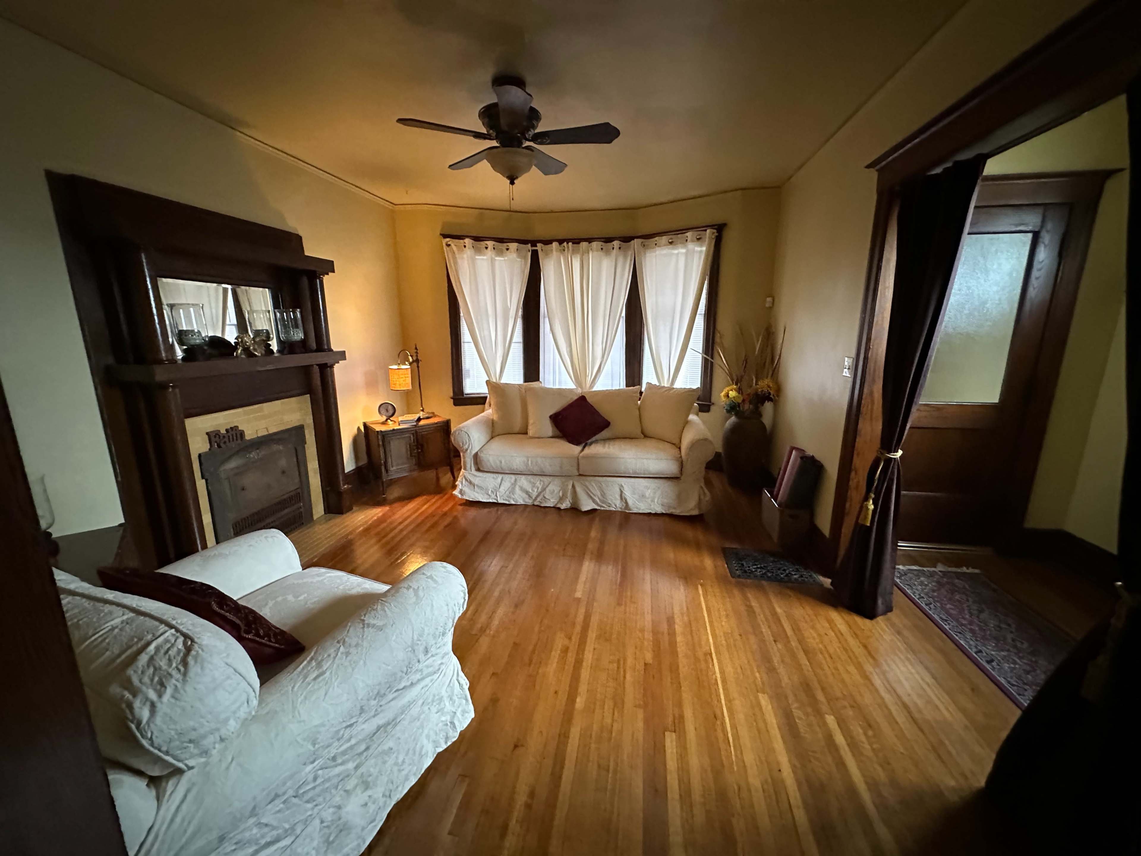 The room features two sofas, a fireplace, and large windows with sheer curtains, all set on polished wooden floors.