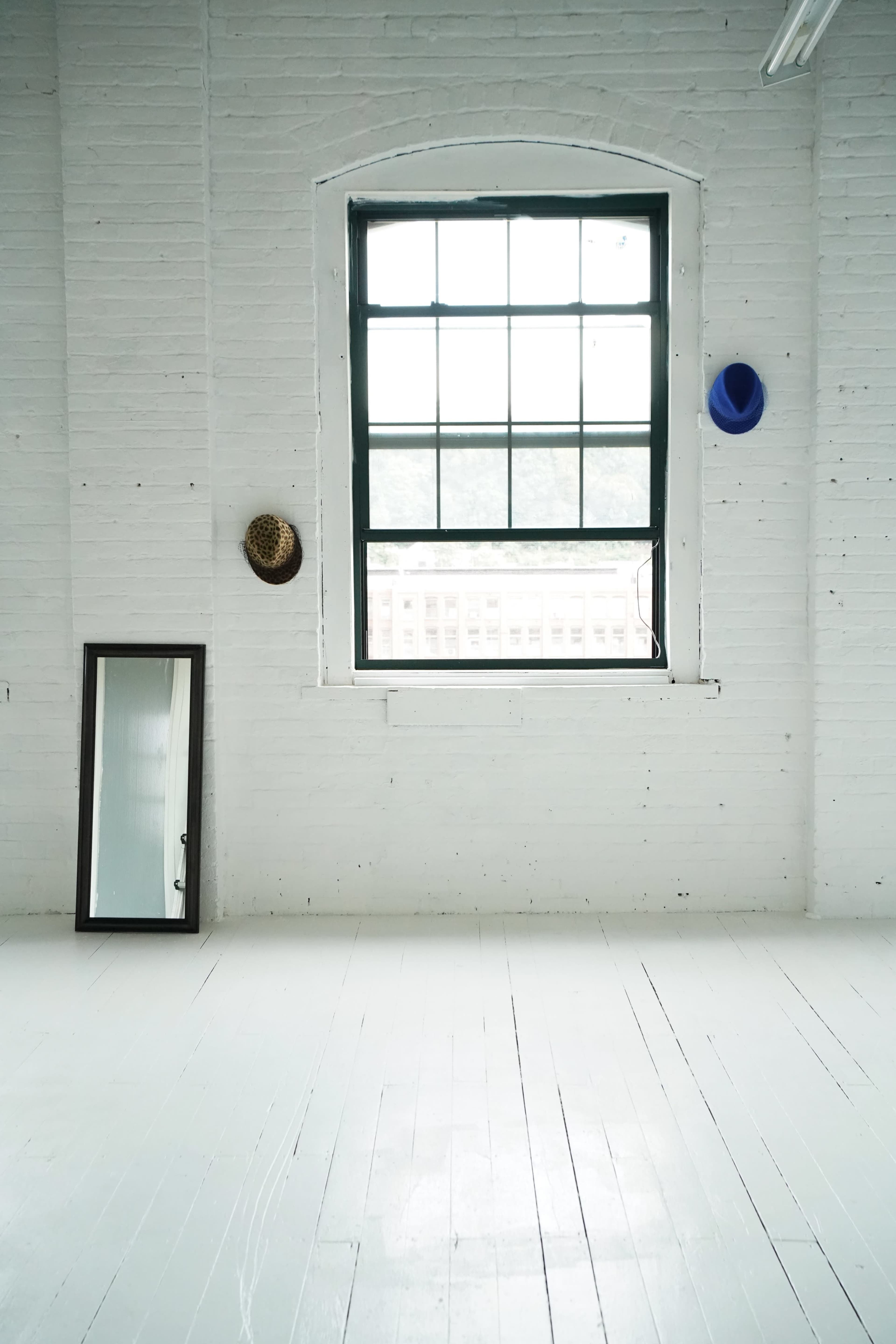 A large window is set in a white brick wall, next to a full-length mirror and two hats hanging on the wall.