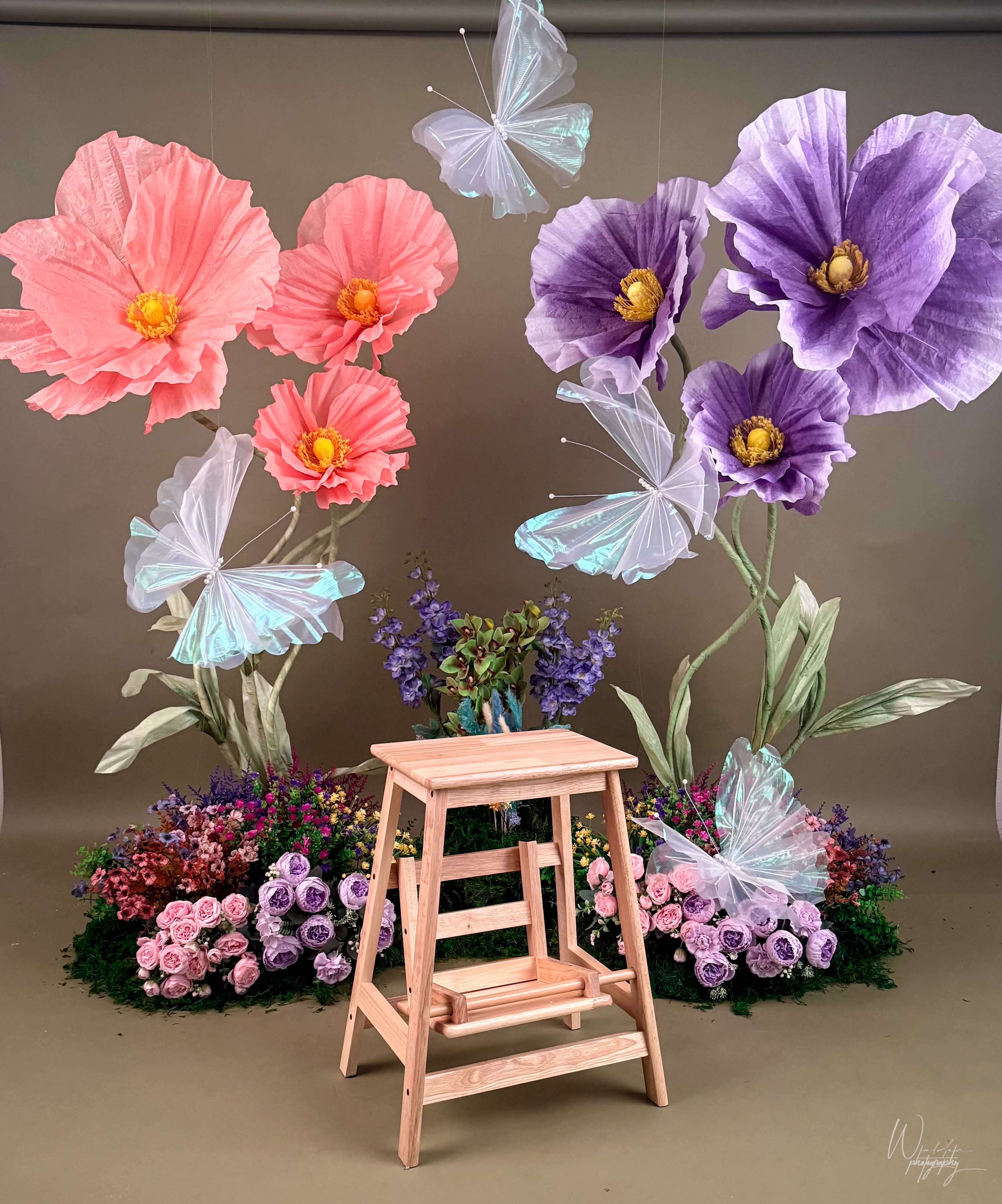 The image features a wooden ladder against a backdrop of large, colorful paper flowers and butterflies, with a floral arrangement at the bottom.