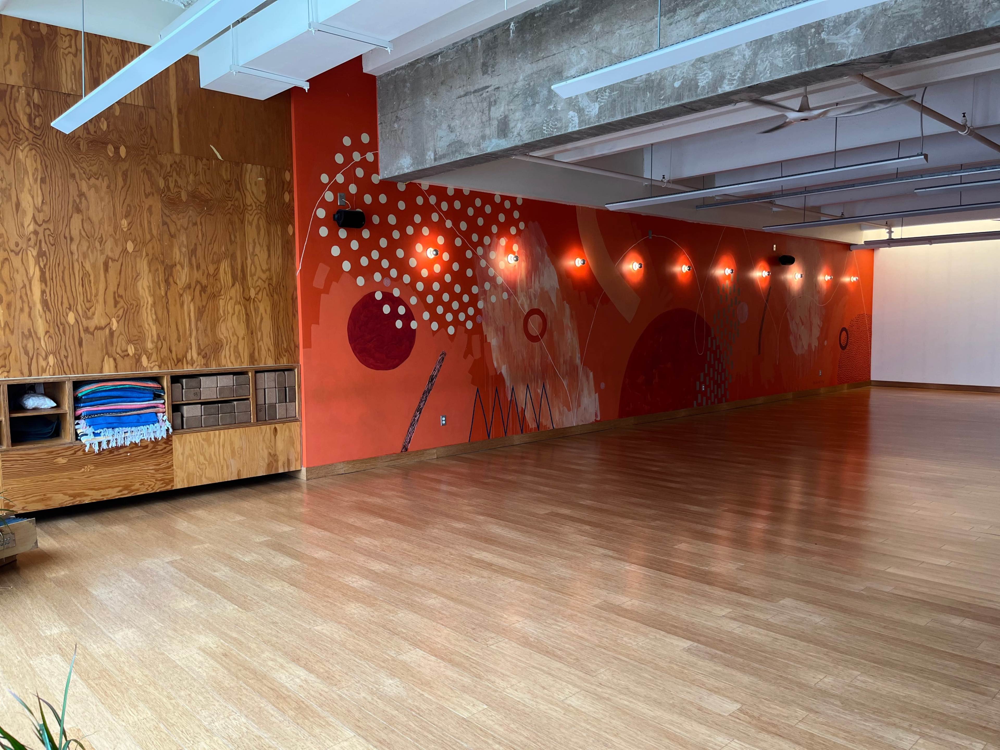 A spacious studio with wooden flooring, featuring a decorative orange wall with abstract patterns and a wooden storage unit on the left.