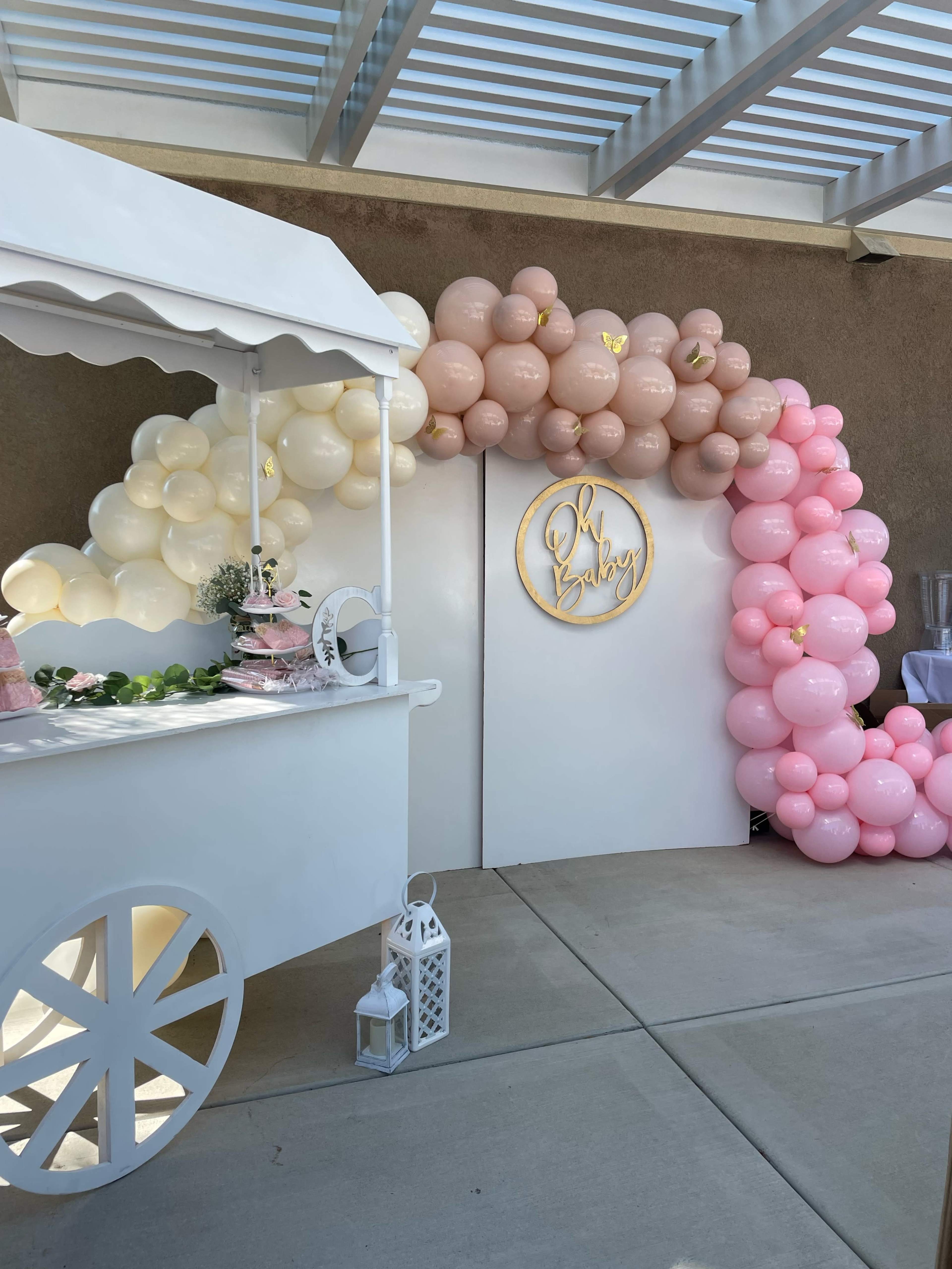 A decorated entrance with a white backdrop and a balloon arch in shades of pink and beige is complemented by a candy cart nearby.